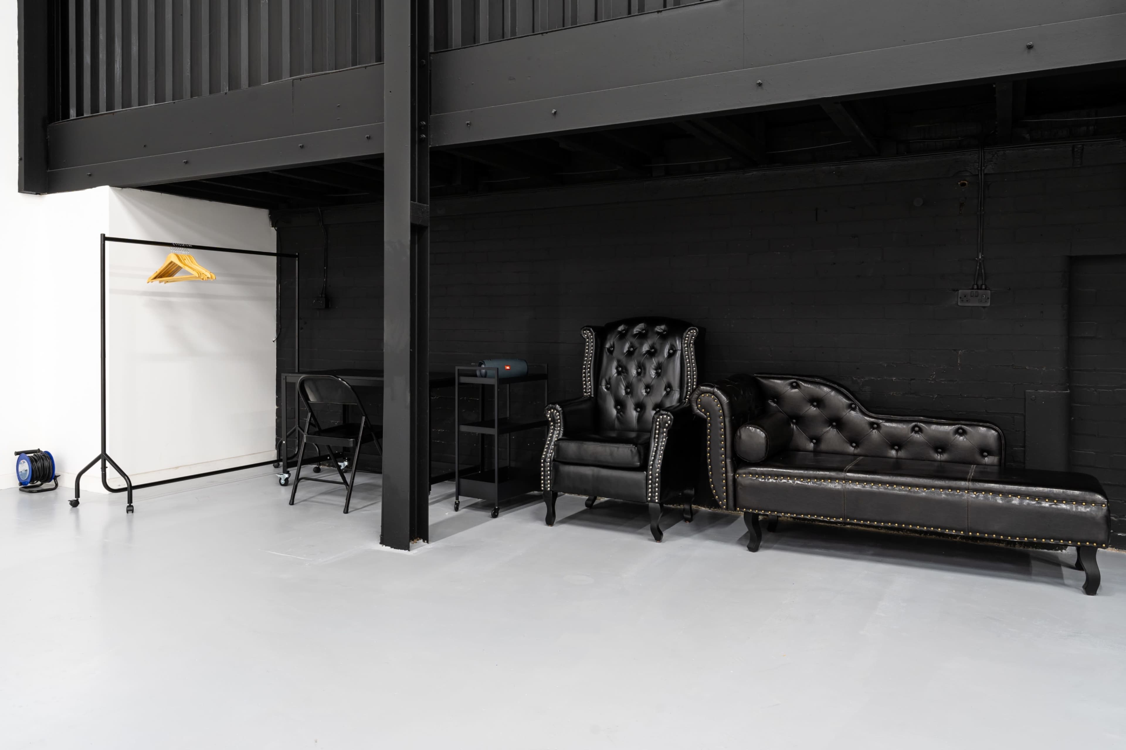 A minimalist interior space with a black leather sofa, a black armchair, a clothing rack, a small table, and a folding chair against a dark wall.