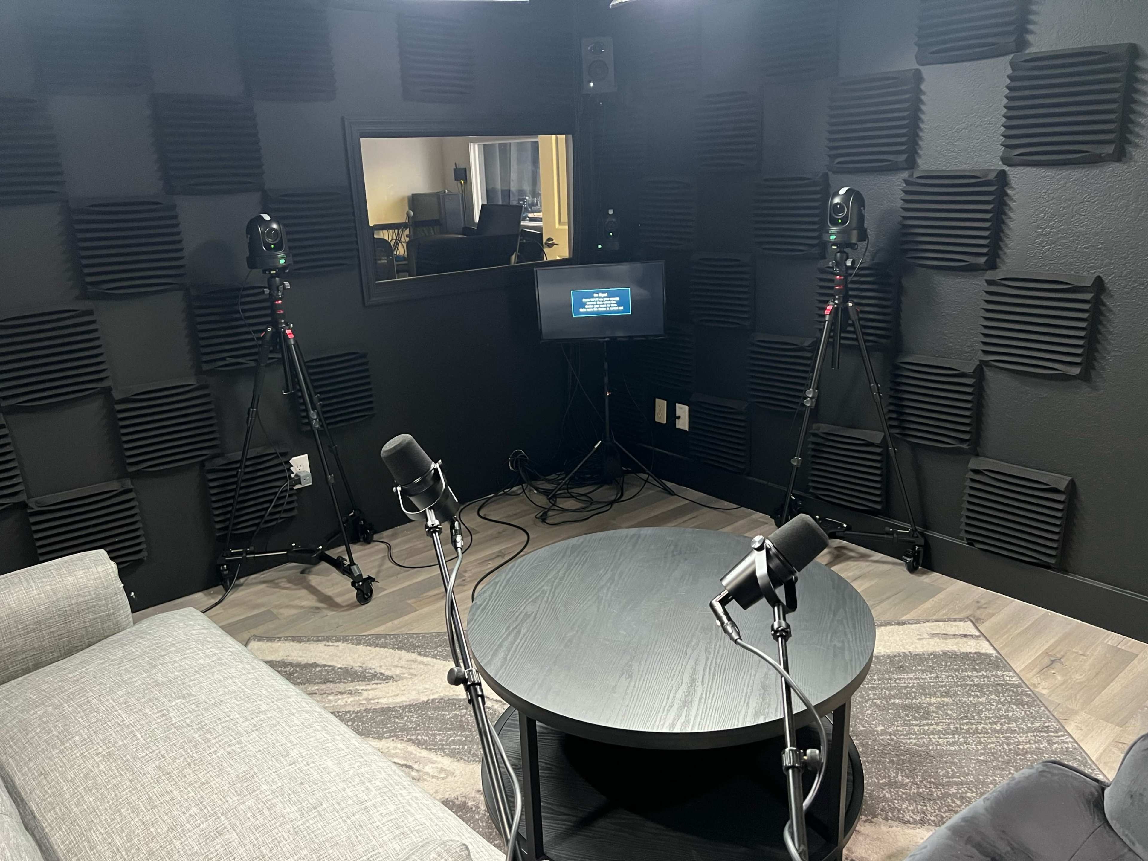 The image shows a small recording studio with soundproofed walls, featuring two microphones on stands, a central table, and cameras positioned around the room.