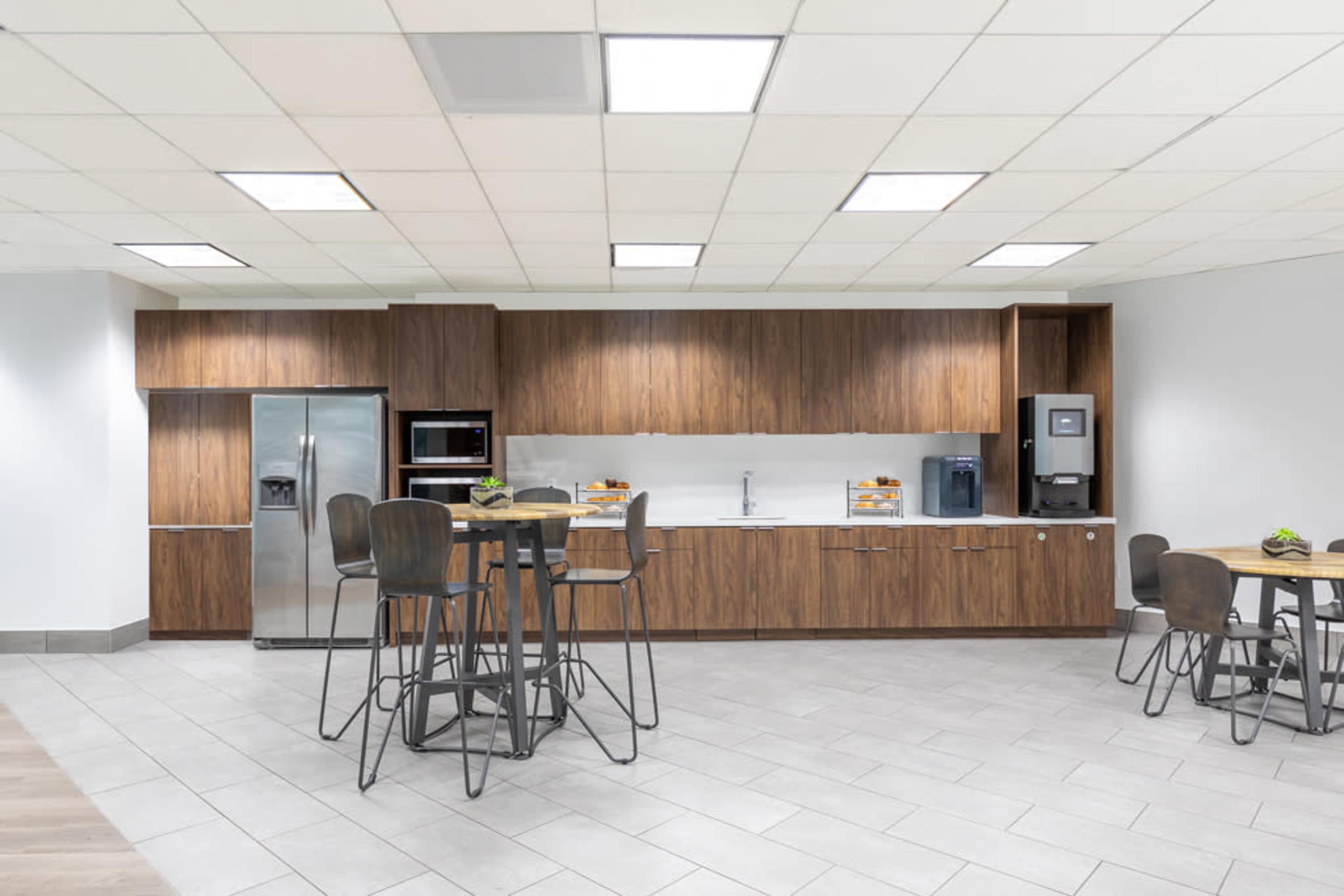 A modern kitchen area featuring brown cabinetry, a refrigerator, a microwave, a coffee machine, and several tall tables with stools.