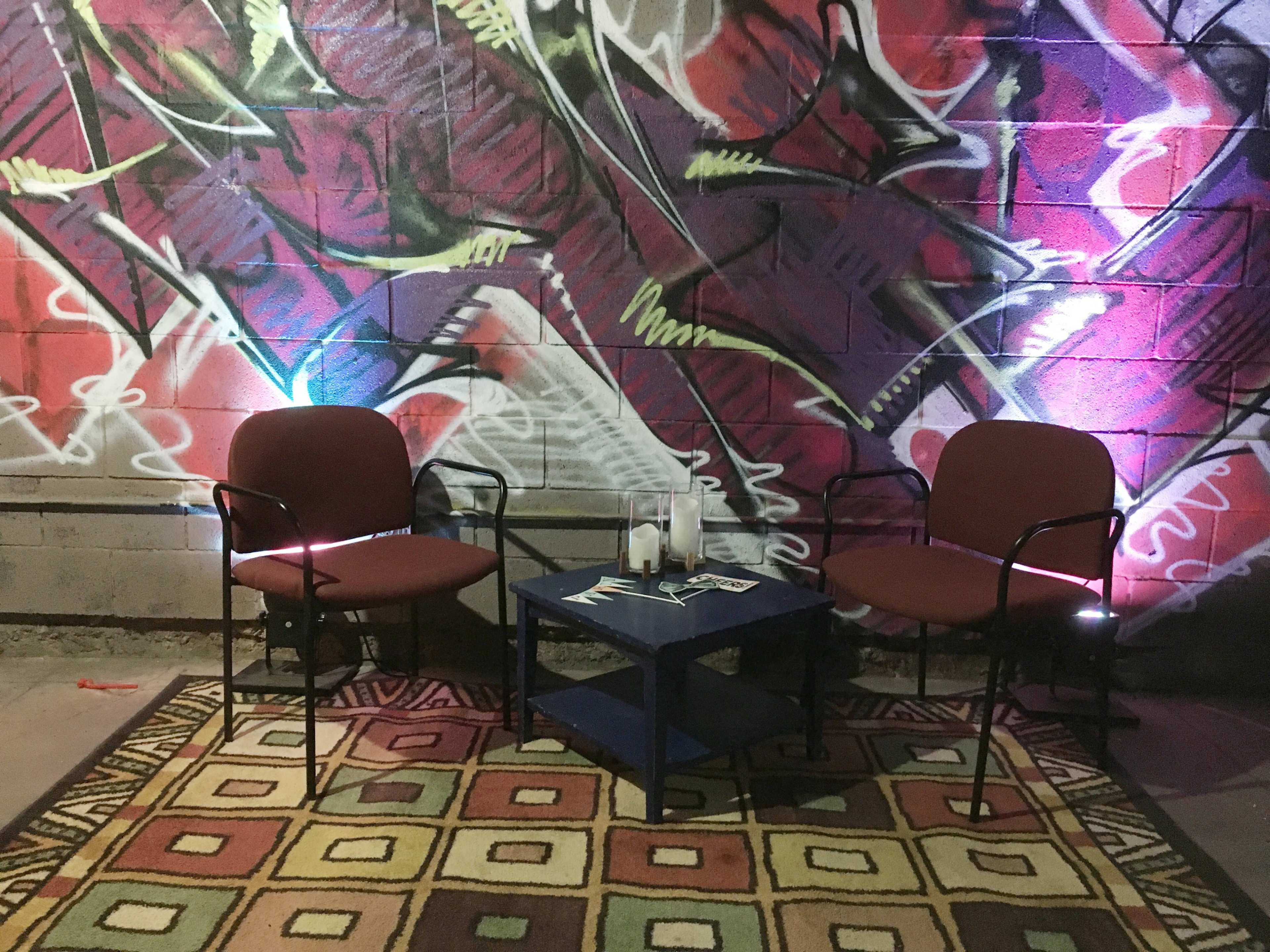 Two red chairs are positioned around a blue table on a patterned rug, with a colorful graffiti mural on the wall behind them.
