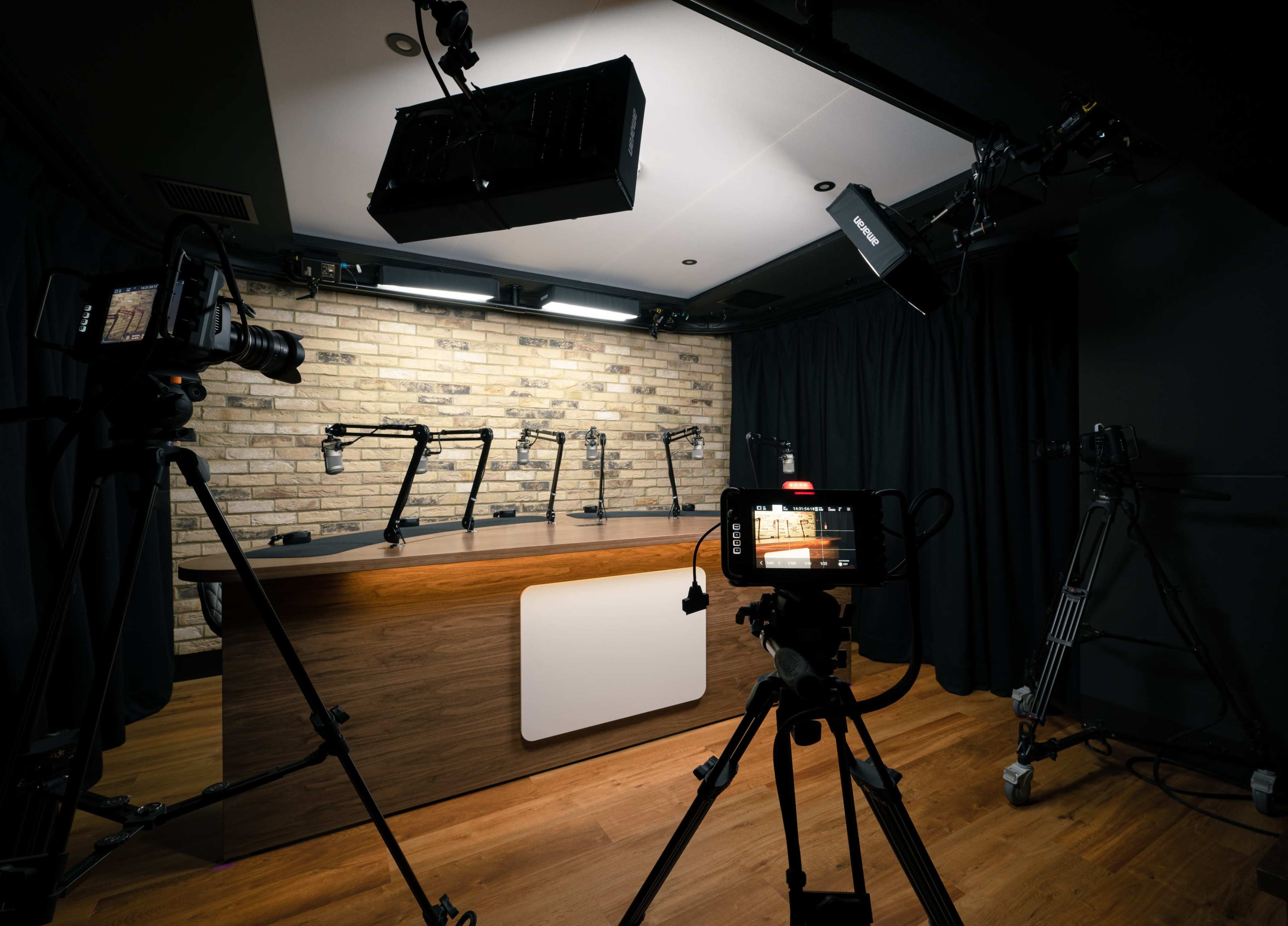 The image shows a video studio setup with multiple cameras positioned in front of a wooden table and a brick wall, illuminated by overhead lights.