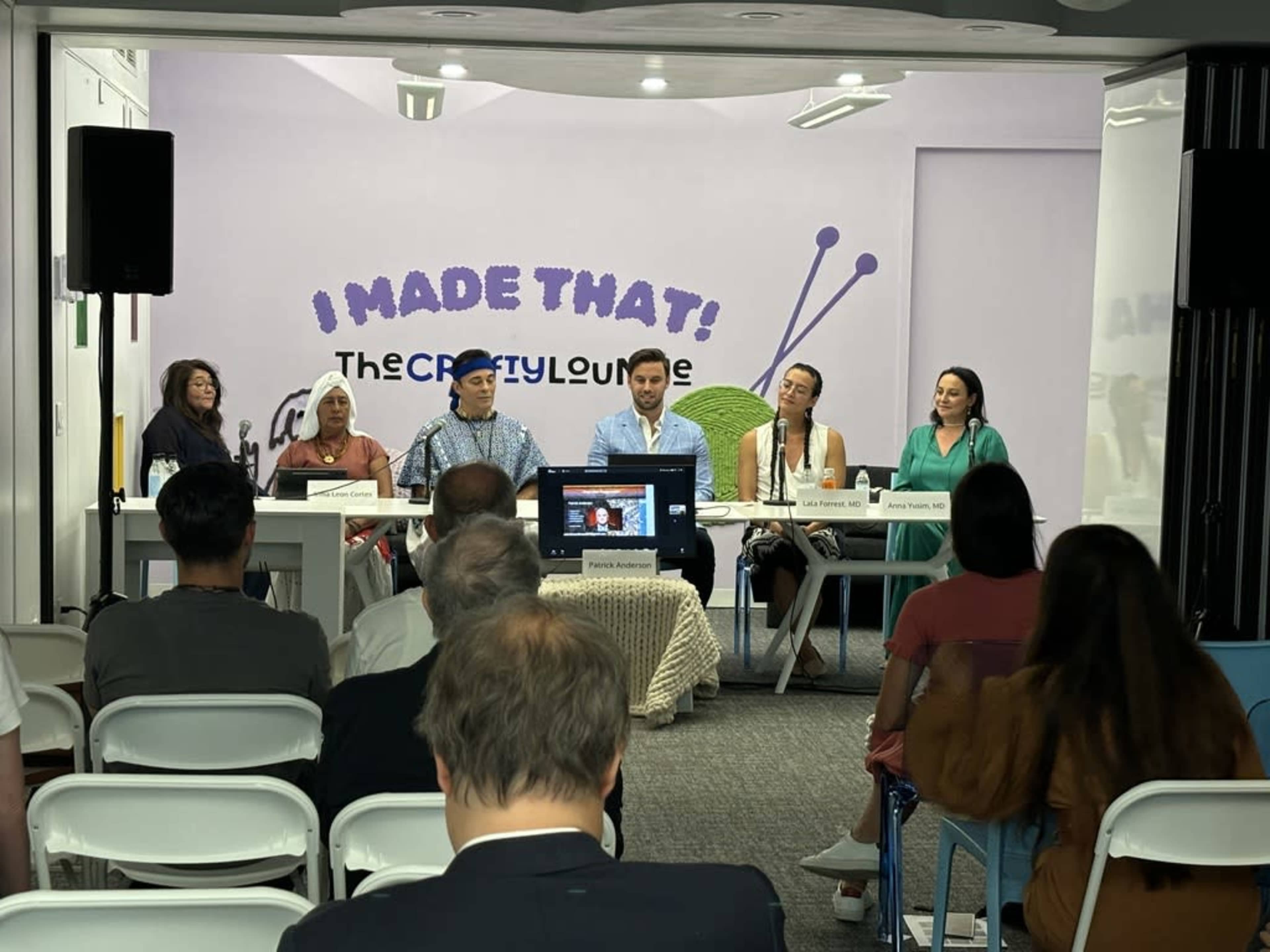A panel discussion is taking place in a brightly lit room, with seven people seated at a table and an audience visible in the foreground.