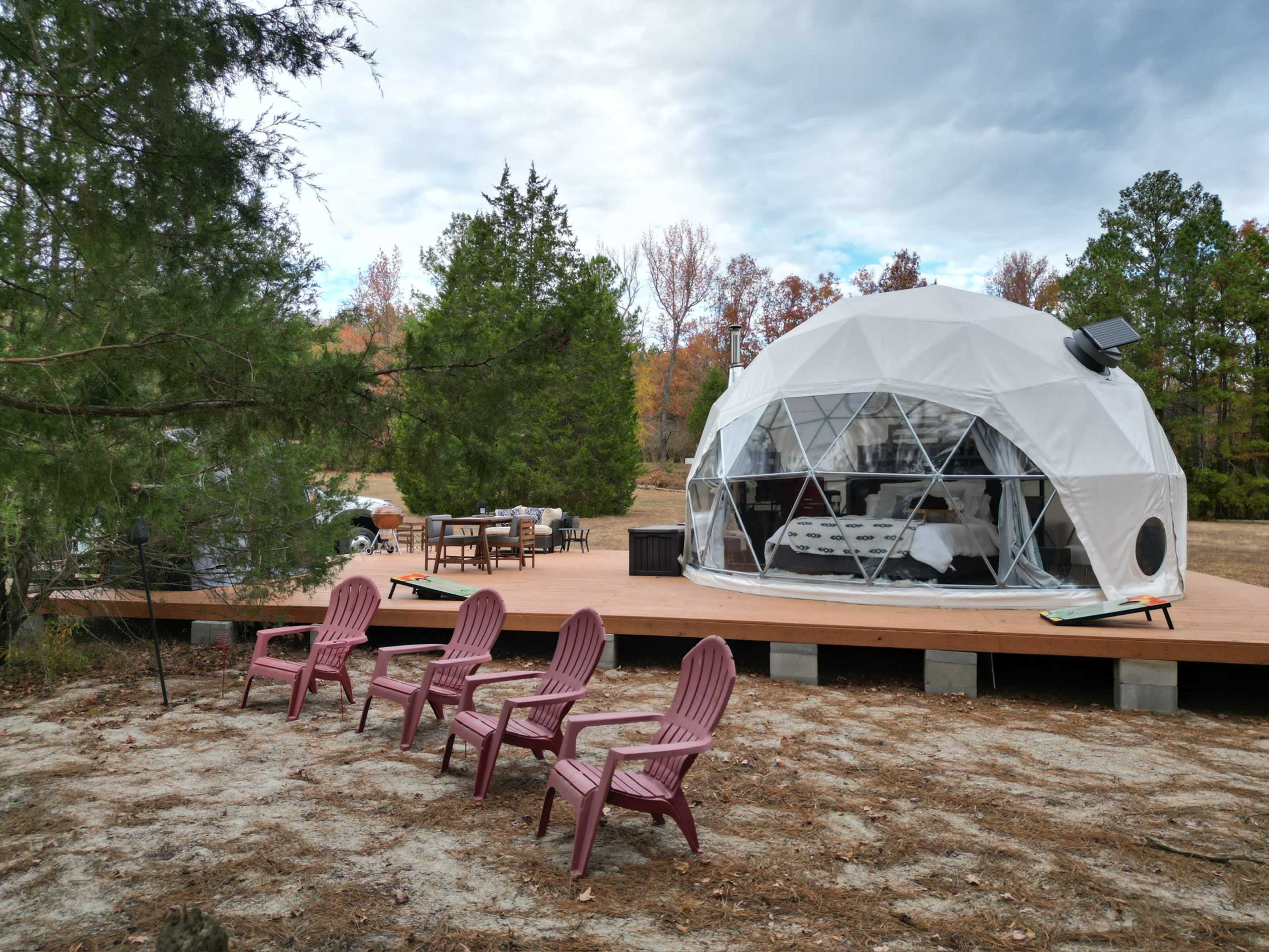 A geodesic dome structure is set on a wooden platform in a wooded area, with several pink Adirondack chairs arranged in front.