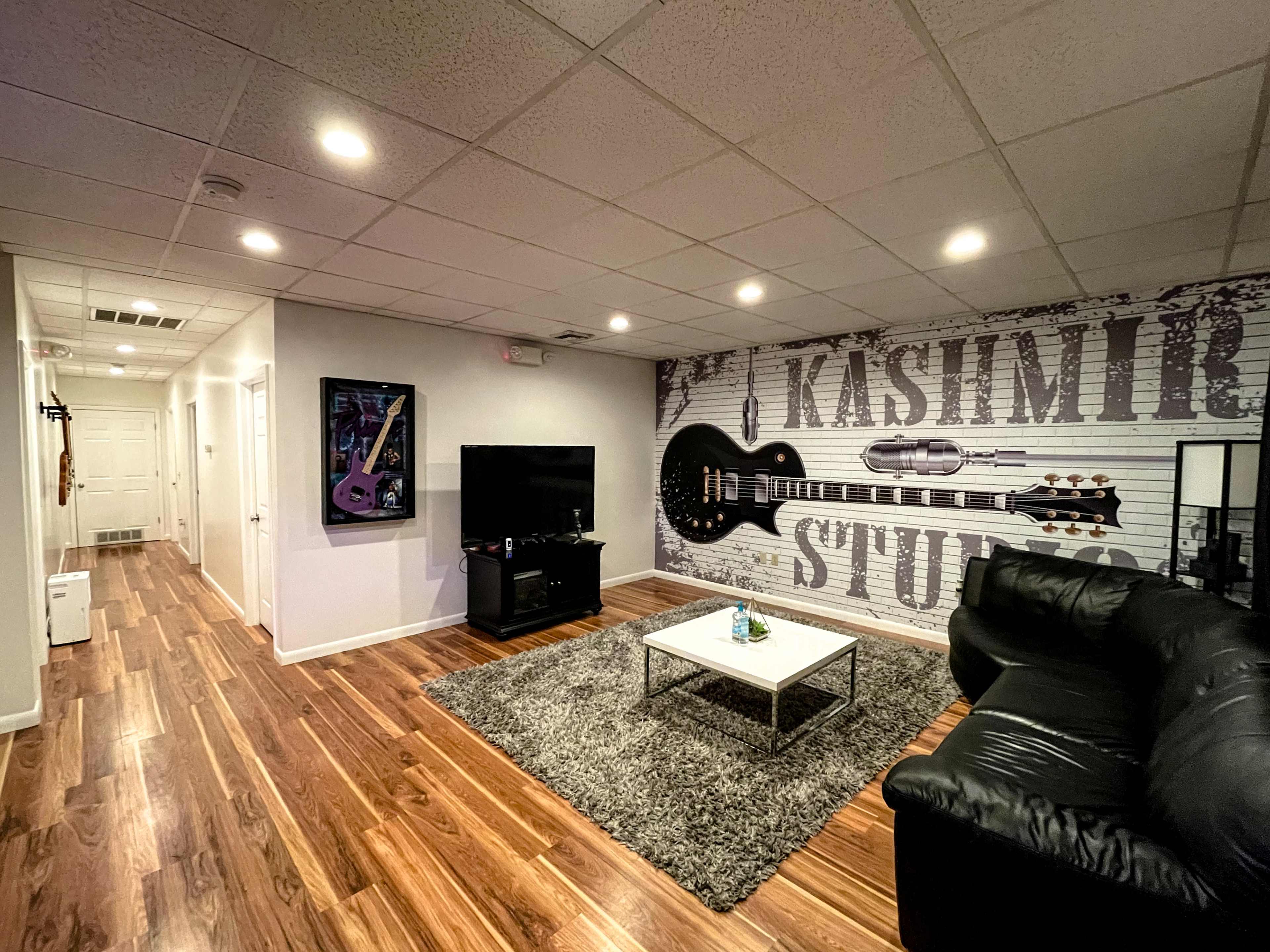 The image shows a living room with a black leather couch, a TV stand, and a wall featuring a guitar-themed mural, alongside a hallway leading to other rooms.