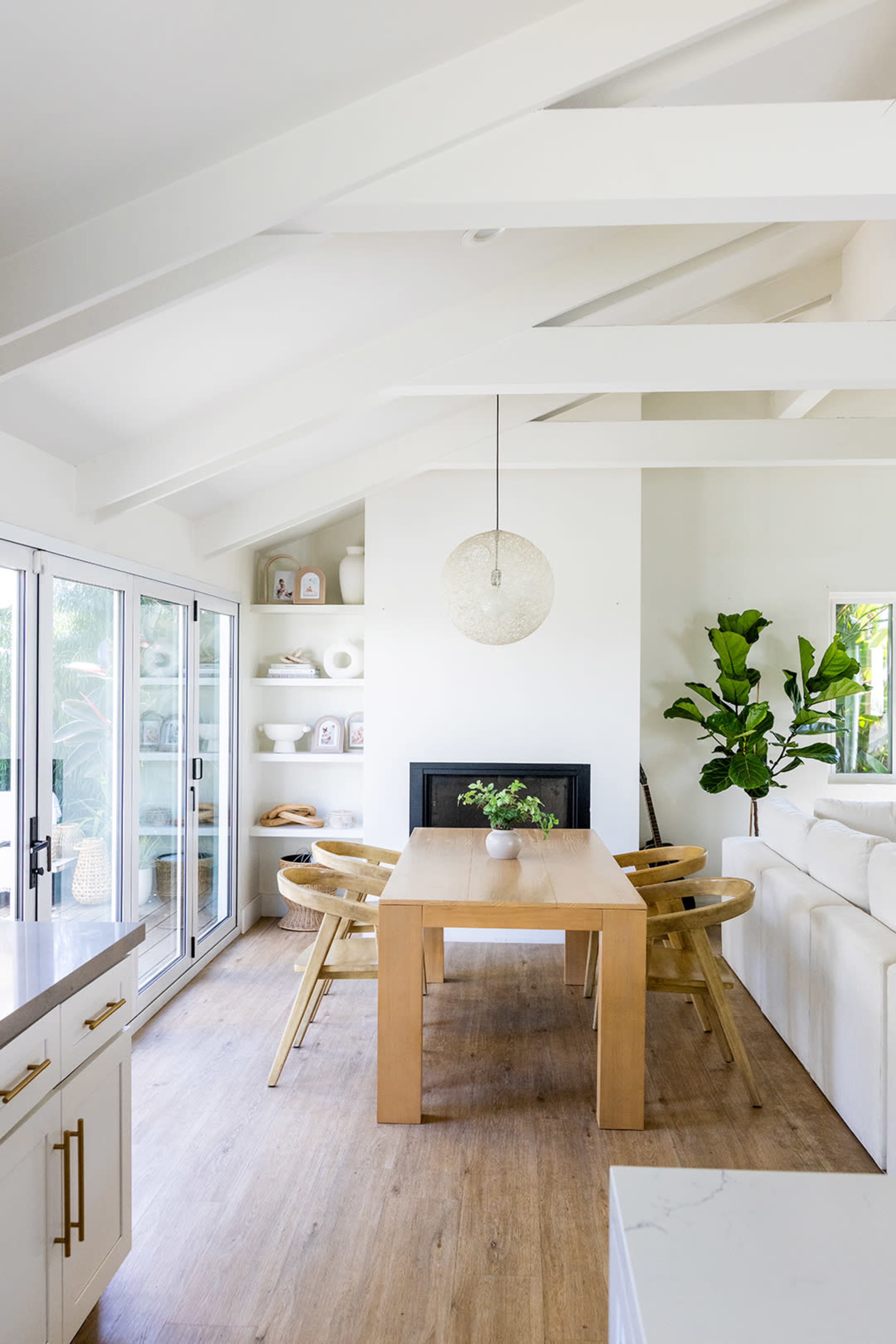 The image shows a modern dining area with a wooden table, four chairs, a large window, a fireplace, and a potted plant.