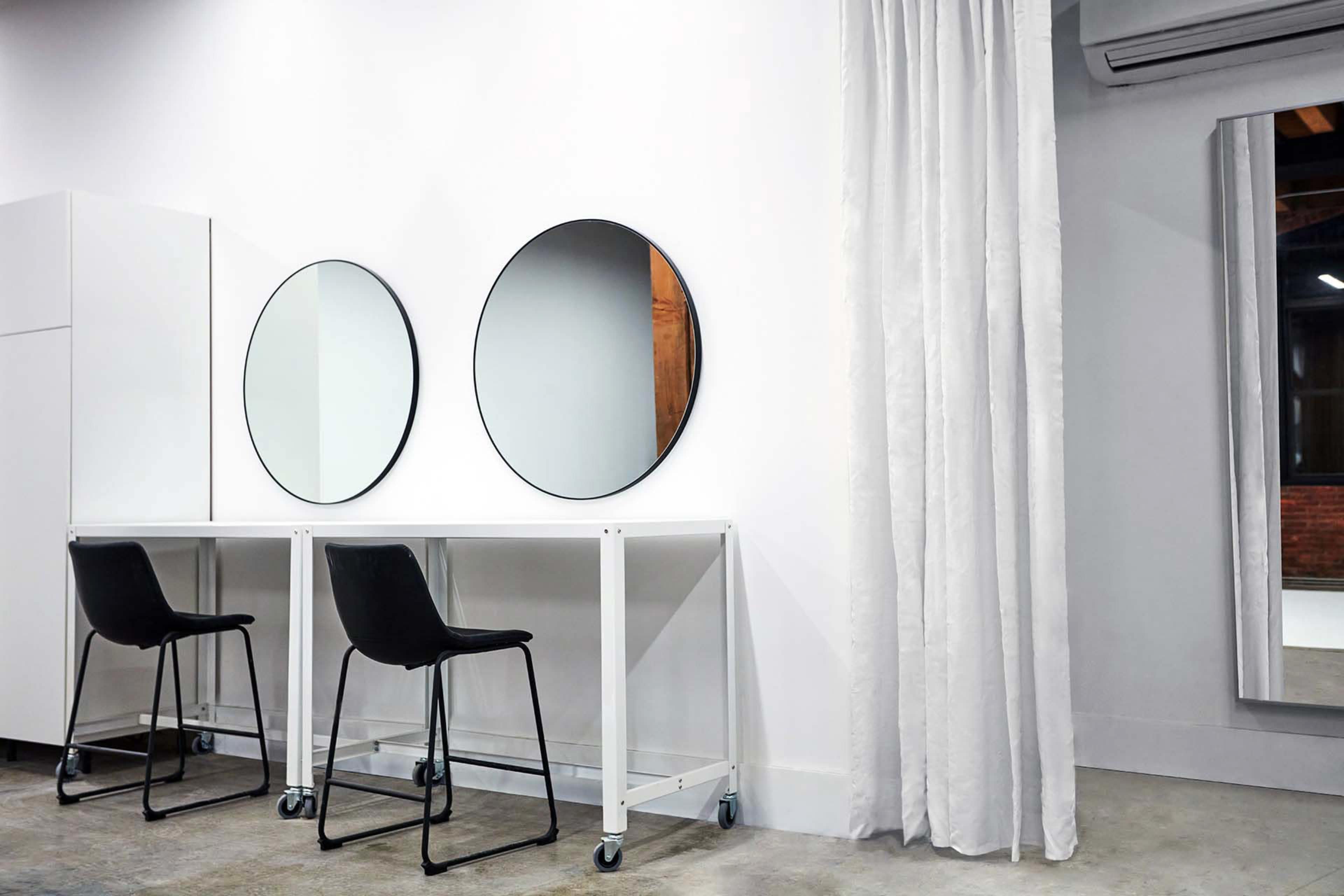 The image shows a minimalist hair salon interior with two circular mirrors above a white table and two black chairs, beside a curtain and a large rectangular mirror on the wall.