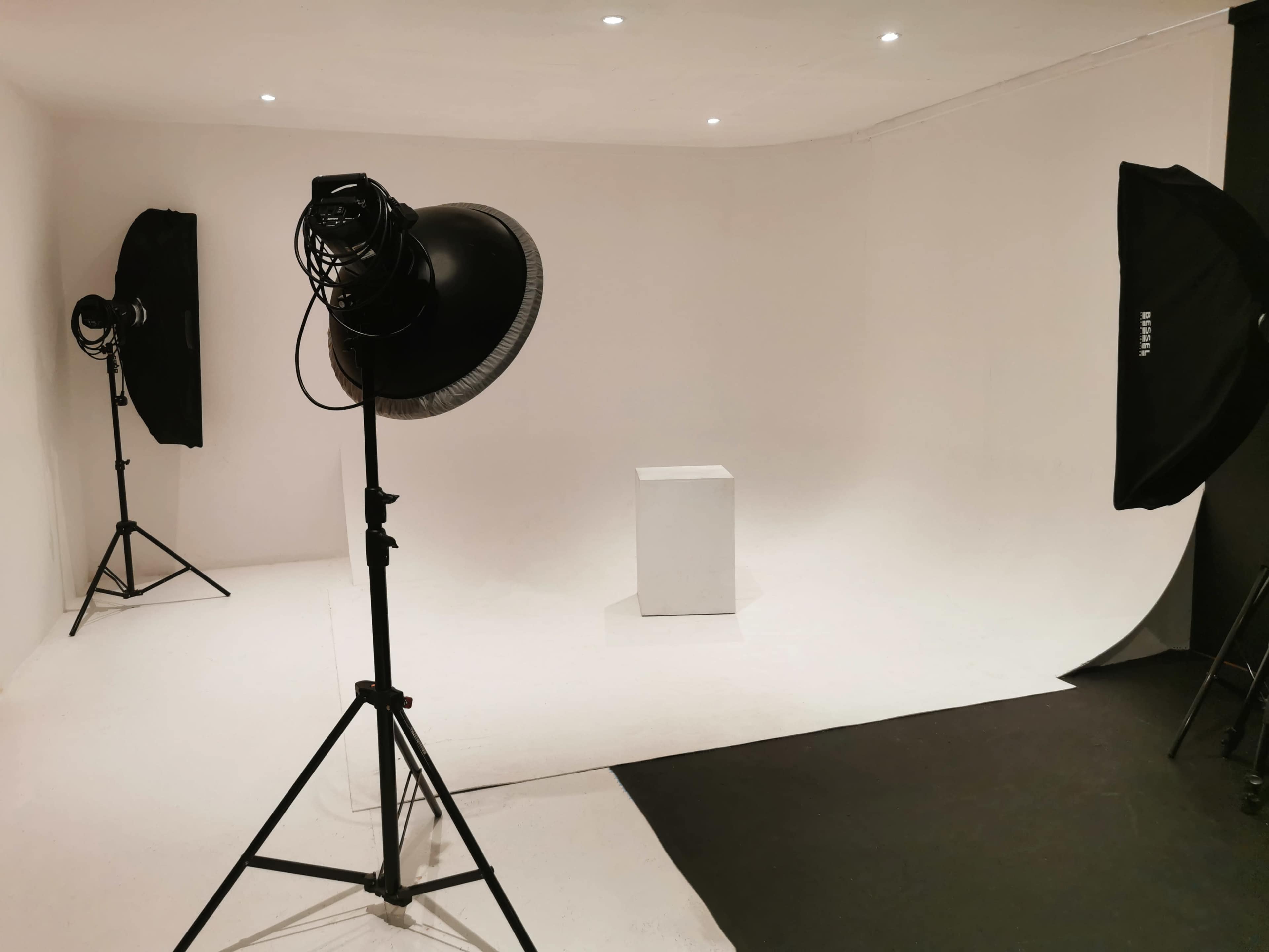 The image shows a photography studio with two large softbox lights and a white backdrop featuring a small white pedestal in the center.