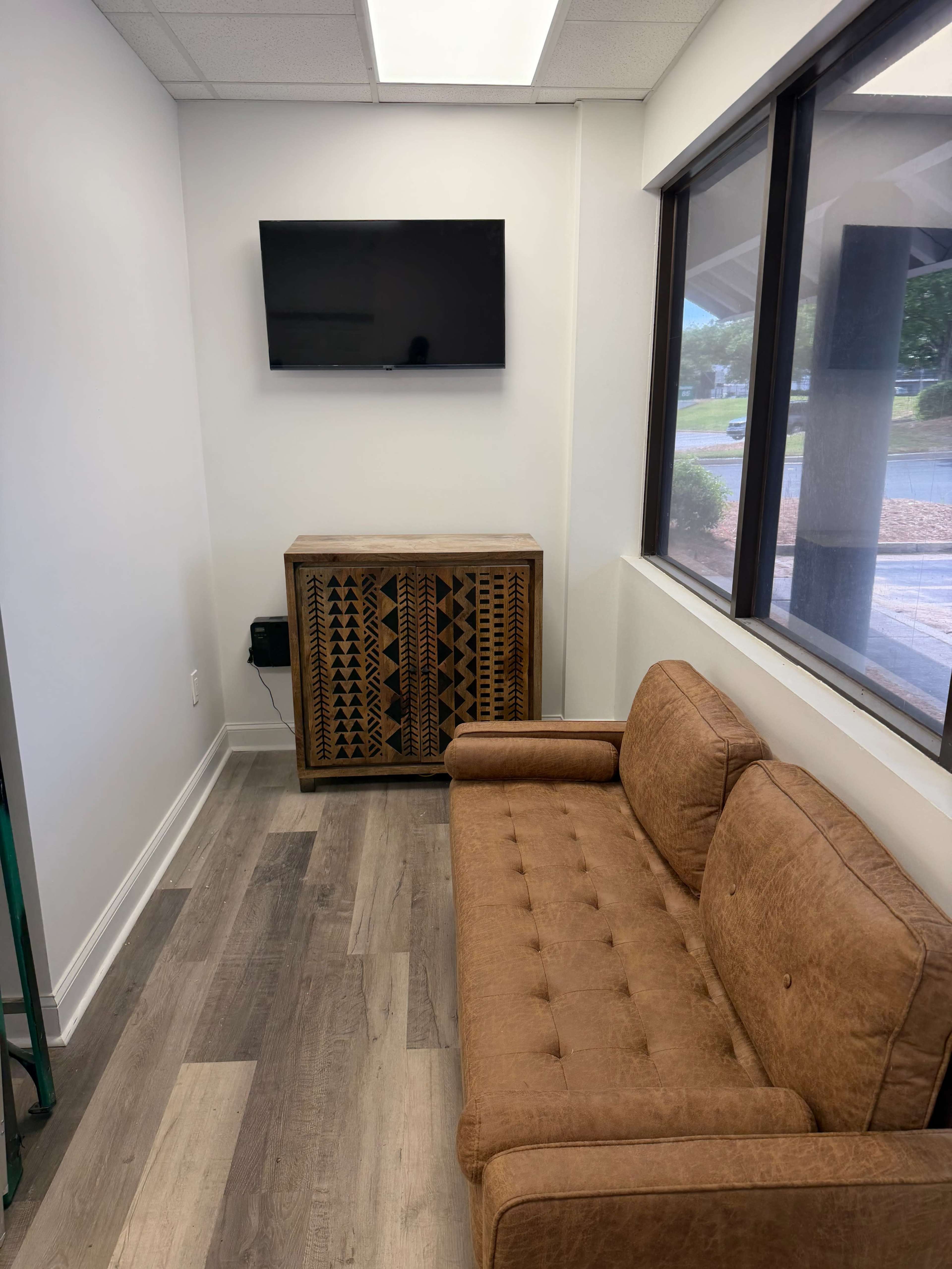 The image shows a small waiting area with a brown couch, a wooden cabinet, and a wall-mounted television.