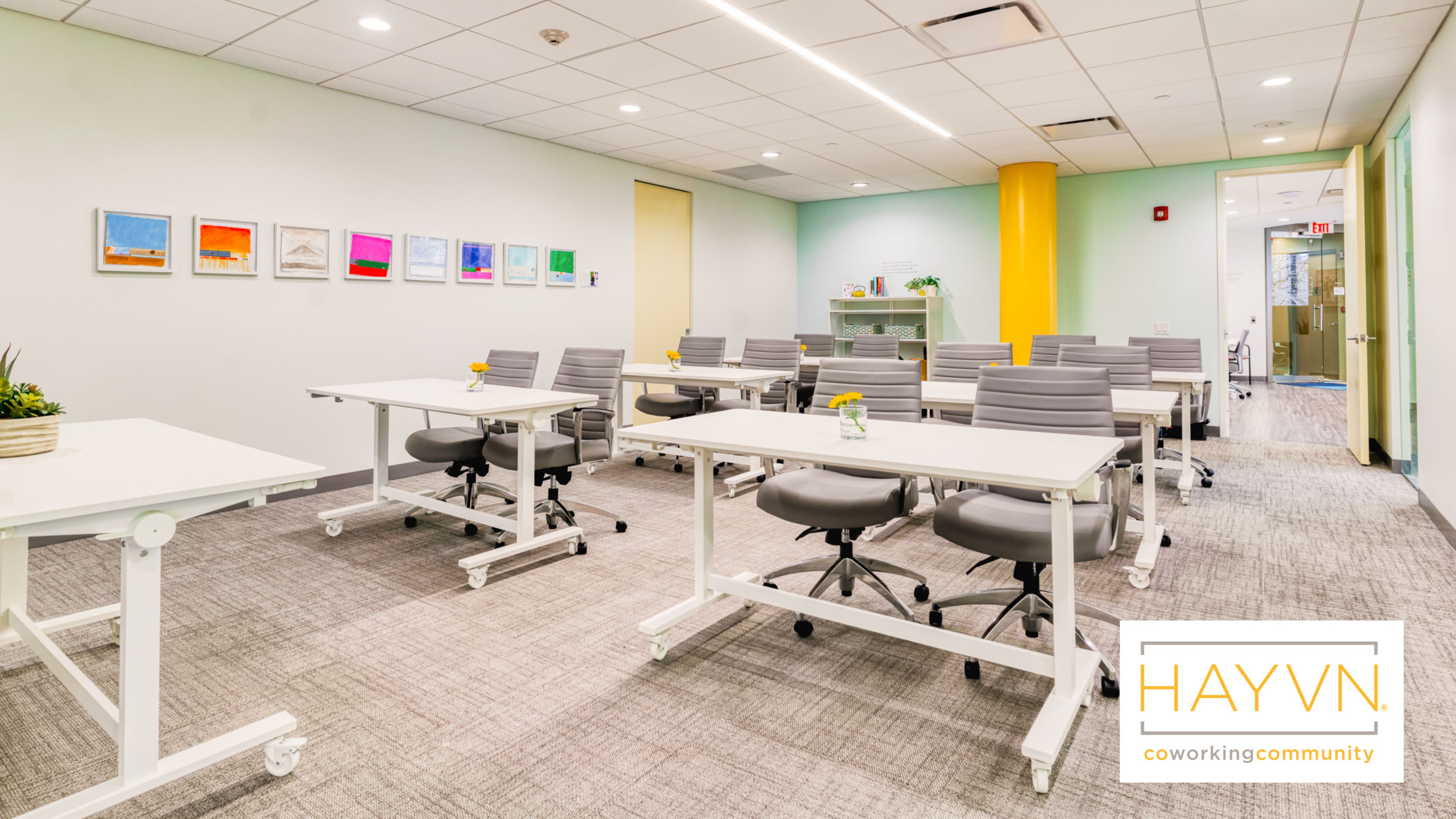 The image shows a bright, modern coworking space with multiple tables and chairs arranged for meetings or classes, and colorful artwork on the wall.