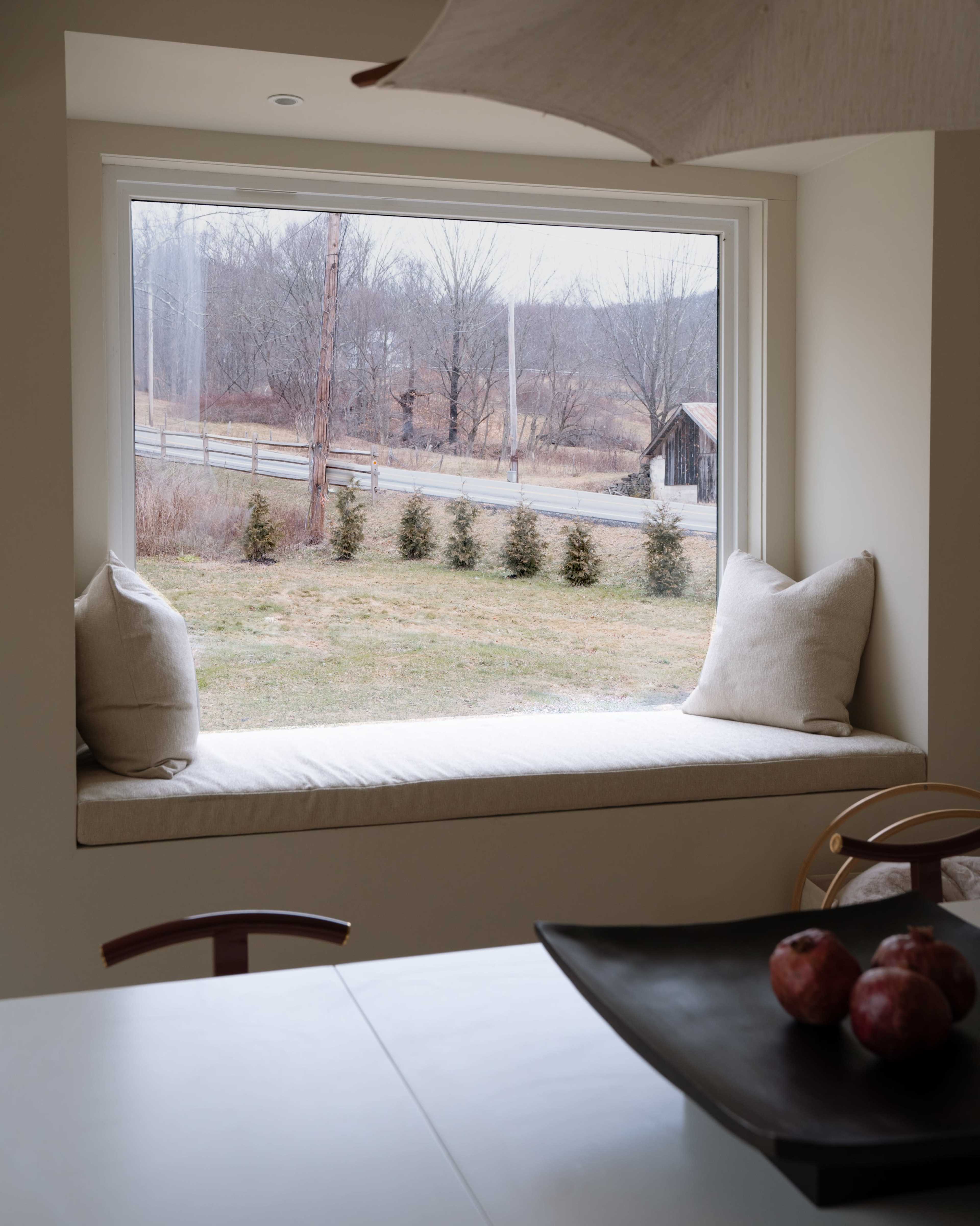A window seat with two pillows overlooking a grassy area and a dirt road lined with small trees.