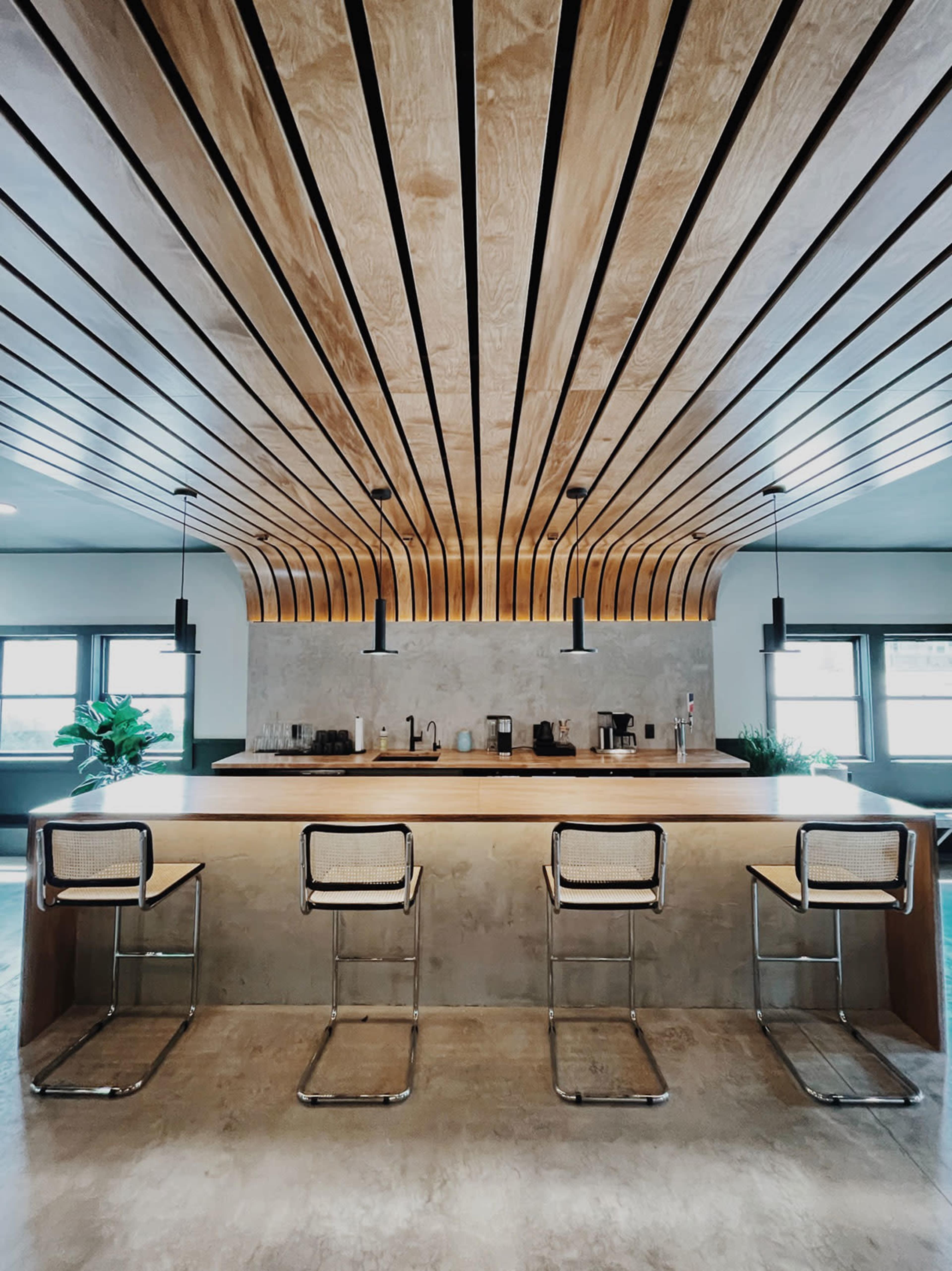A modern kitchen with a wooden slatted ceiling, a concrete countertop, and four metal bar stools.