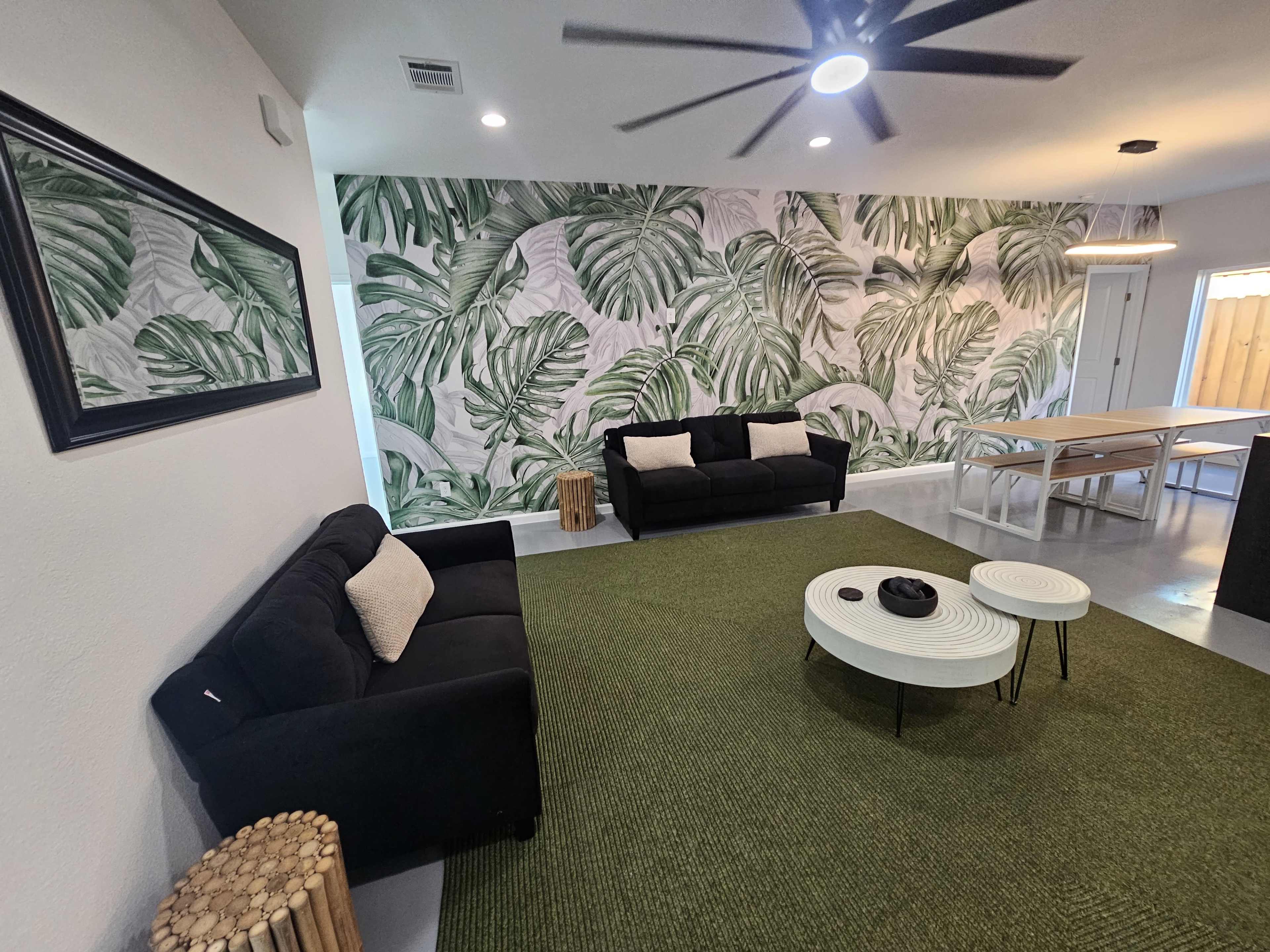 A modern living space with two black sofas, a green area rug, a large wall covered in tropical leaf wallpaper, and a wooden table.