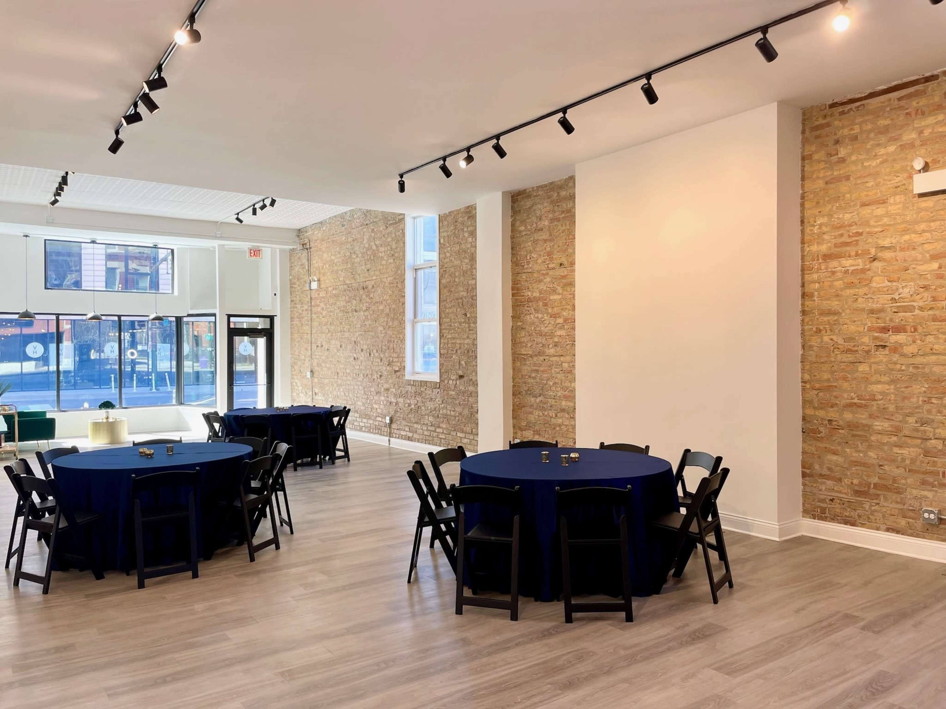 An interior space with two round tables covered in blue tablecloths, black chairs, and exposed brick walls.