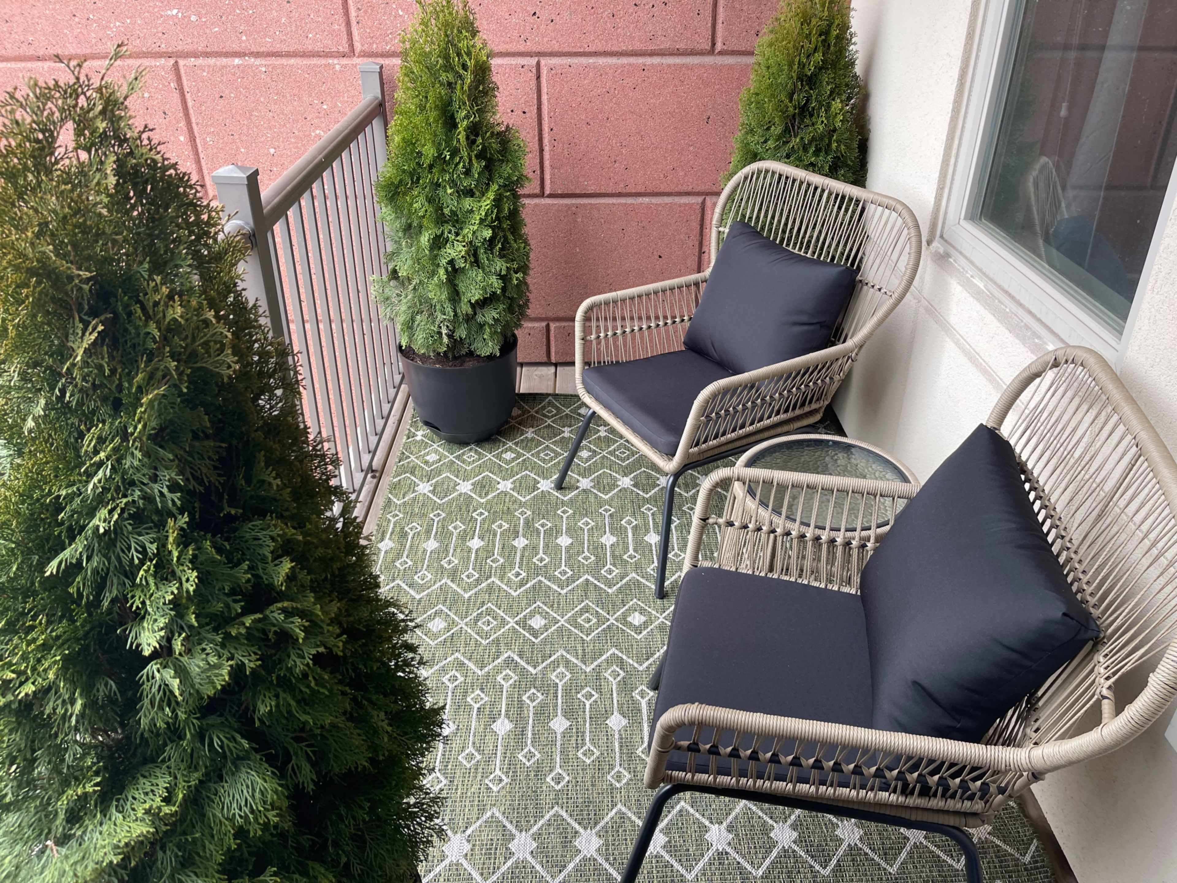 A small balcony featuring two wicker chairs with dark cushions and two potted evergreen plants on either side.