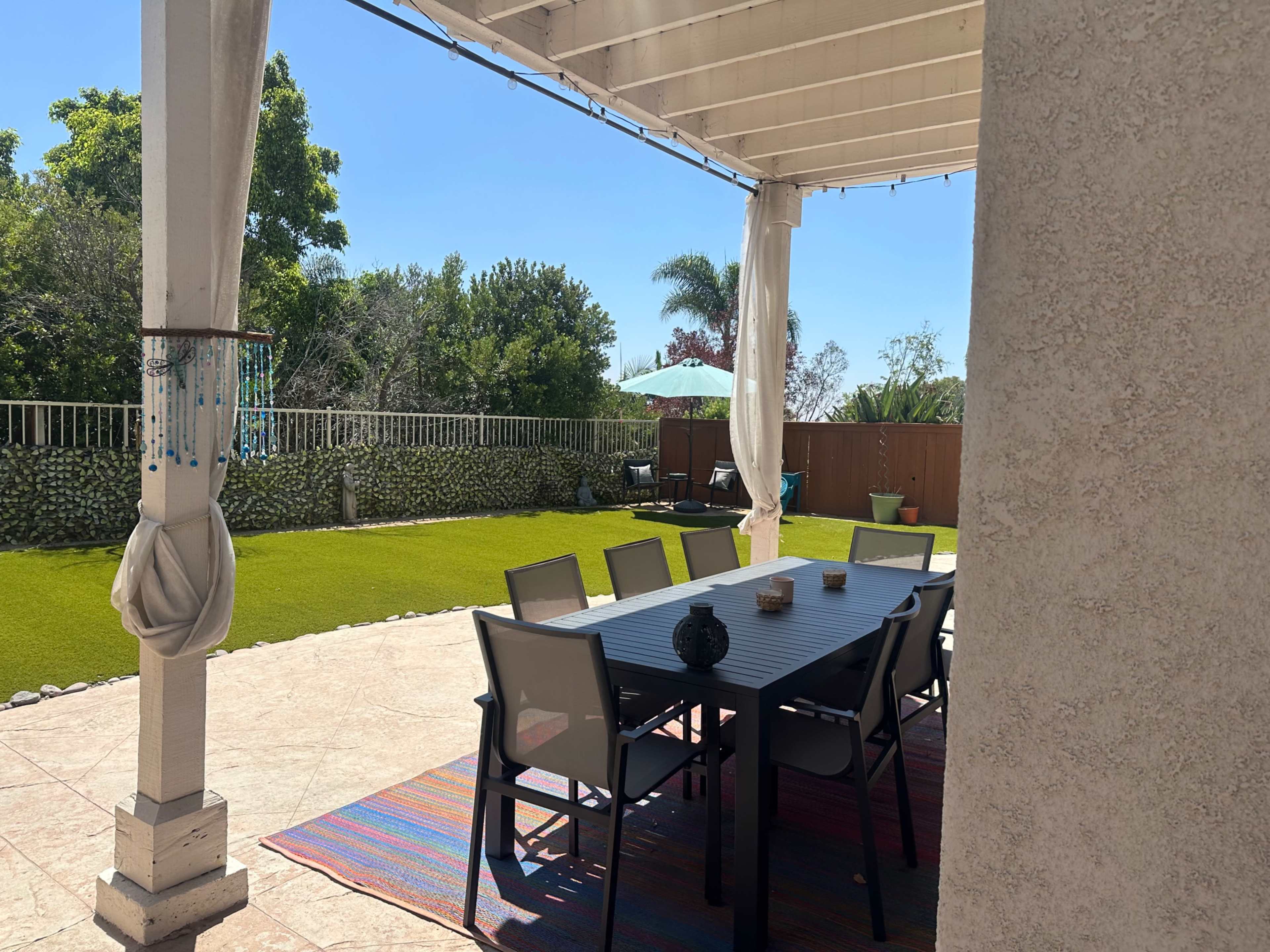 A patio with a dining table and chairs, surrounded by a green lawn and various plants under a clear blue sky.