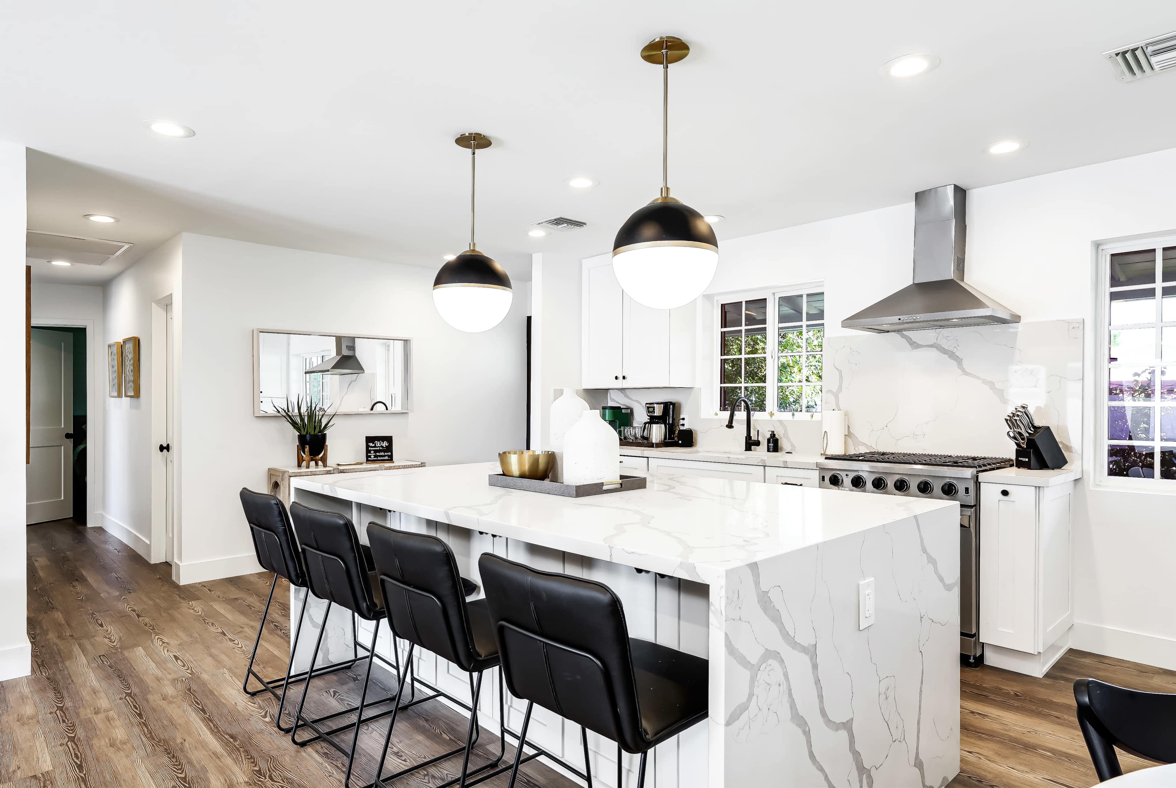 A modern kitchen with a large marble island, black bar stools, pendant lights, and stainless steel appliances.