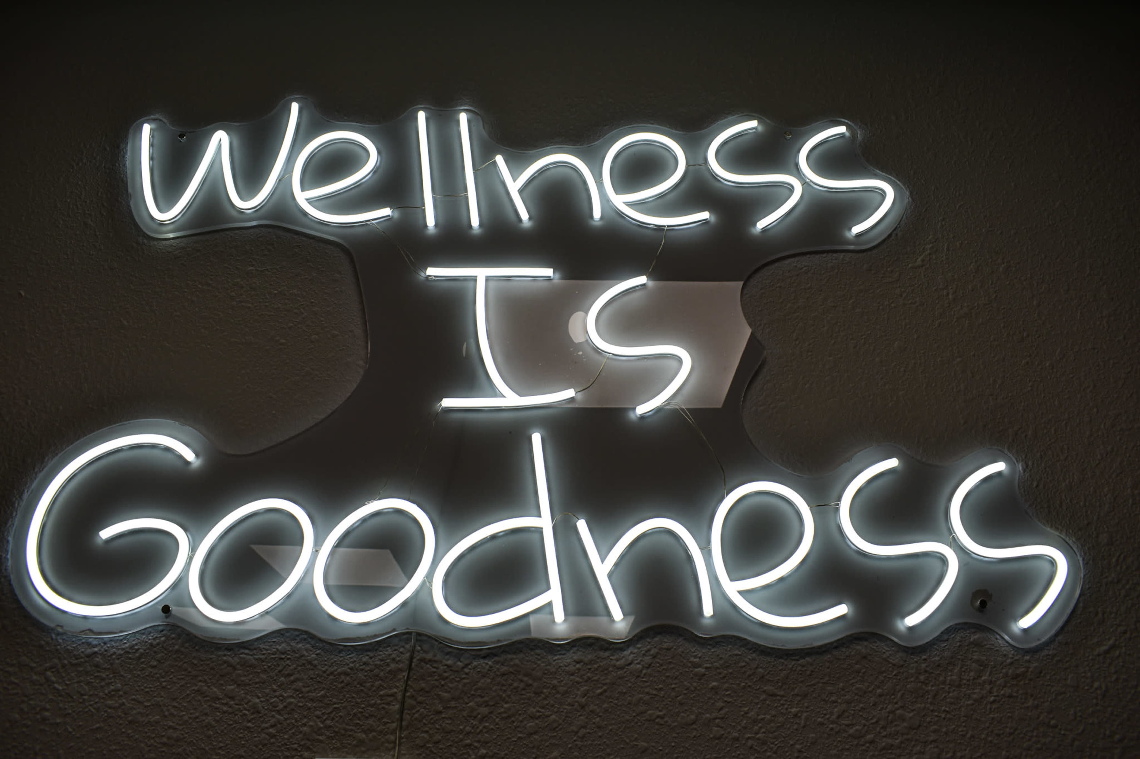 A neon sign reads "Wellness Is Goodness" against a wall.