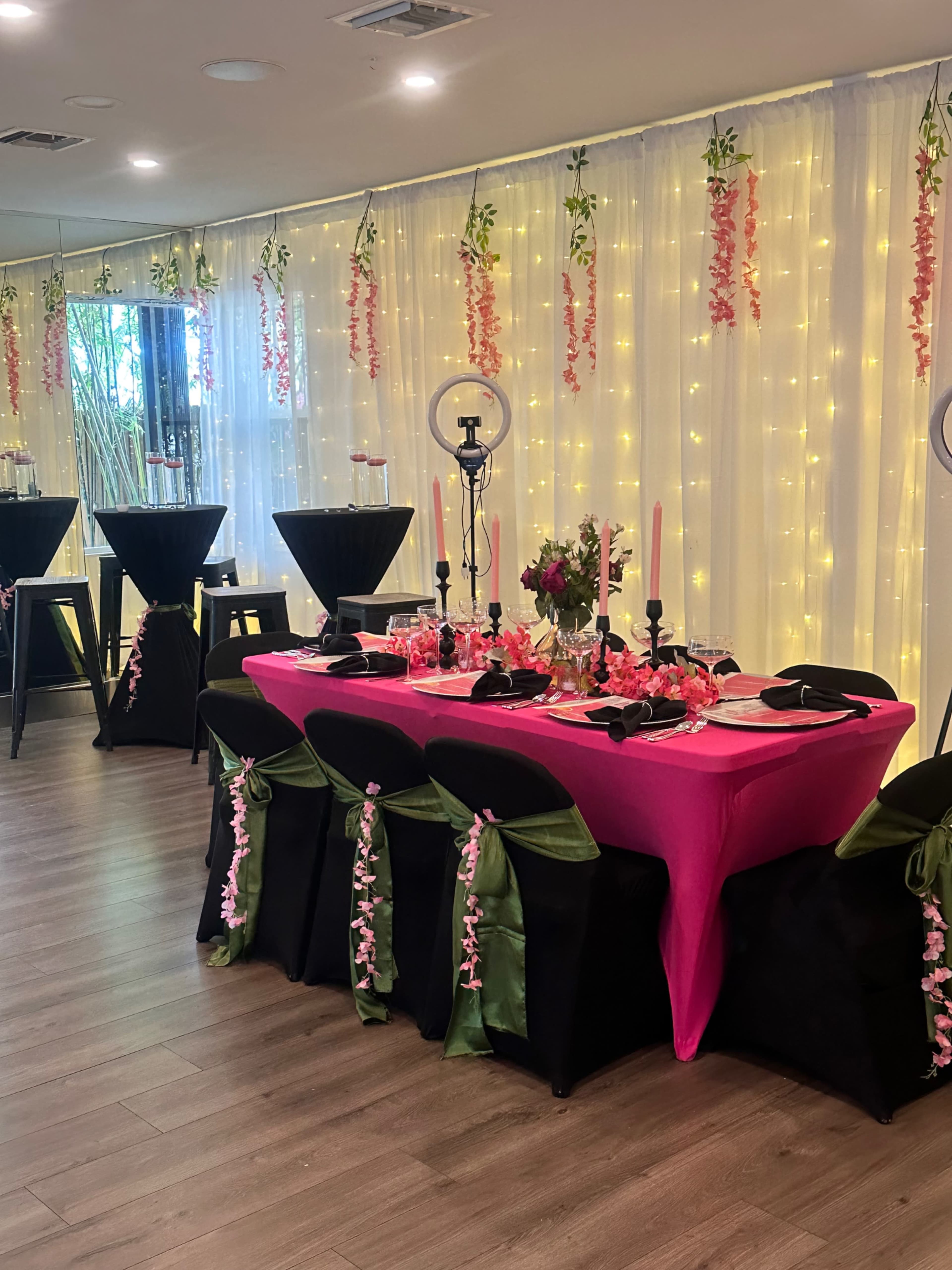 The image shows a decorated dining area with a pink table set for guests, surrounded by black tables and draped lights in the background.