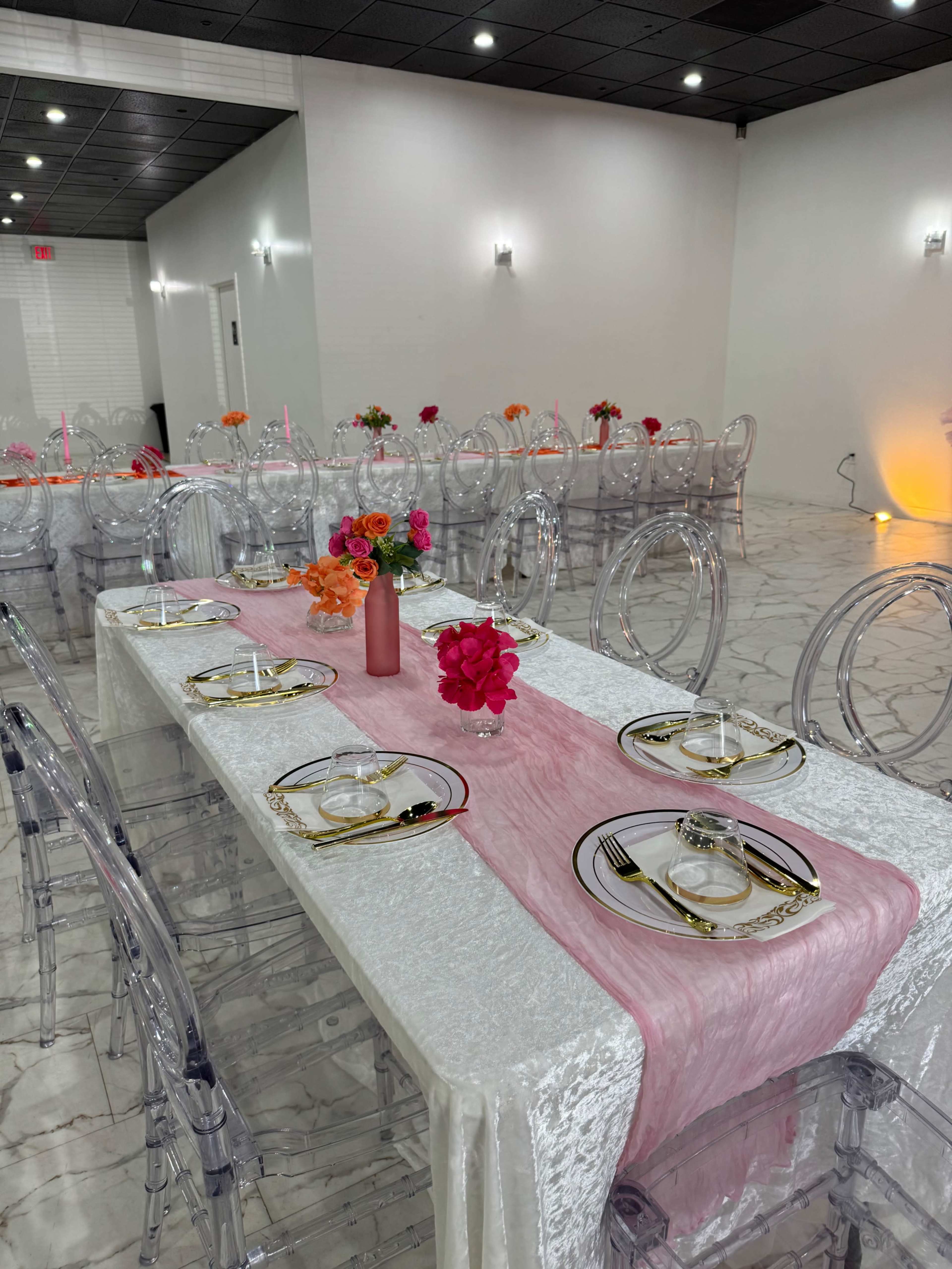 A spacious event hall features long dining tables set with elegant tableware, pink table runners, and colorful floral centerpieces.