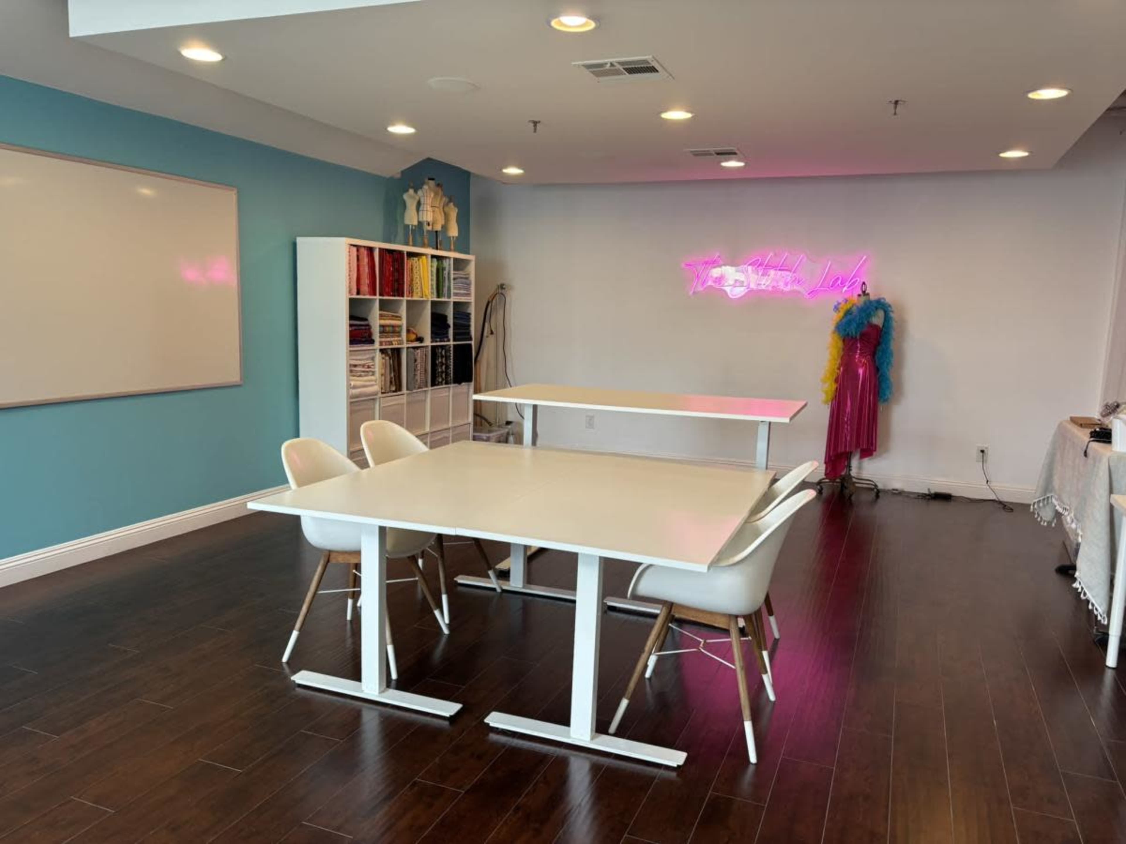A modern, brightly lit room features two white tables and several white chairs against a teal wall, with shelves filled with colorful materials and a neon sign in the background.