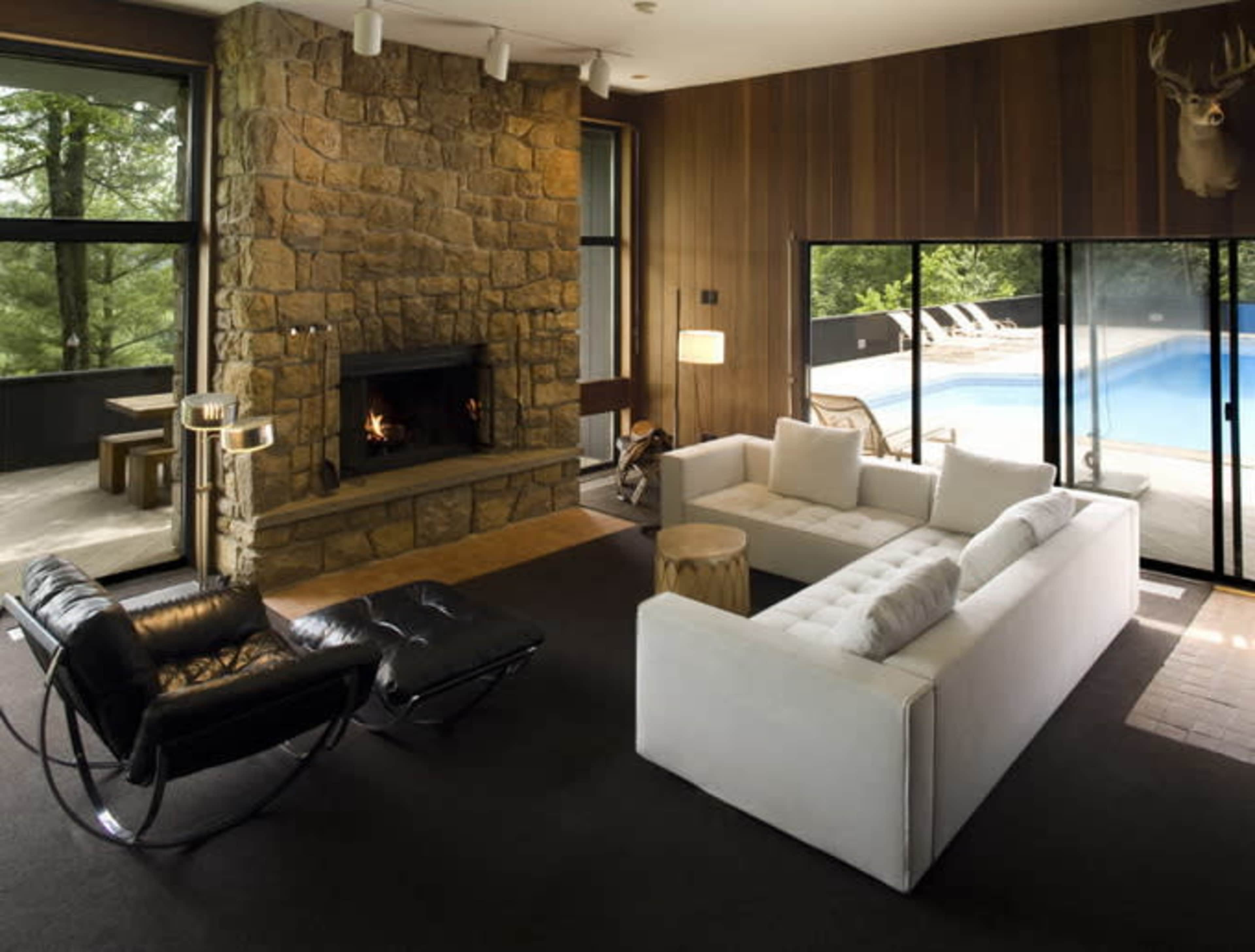 A modern living room features a stone fireplace, a black lounge chair, and a white sectional sofa, with large windows overlooking a pool area.