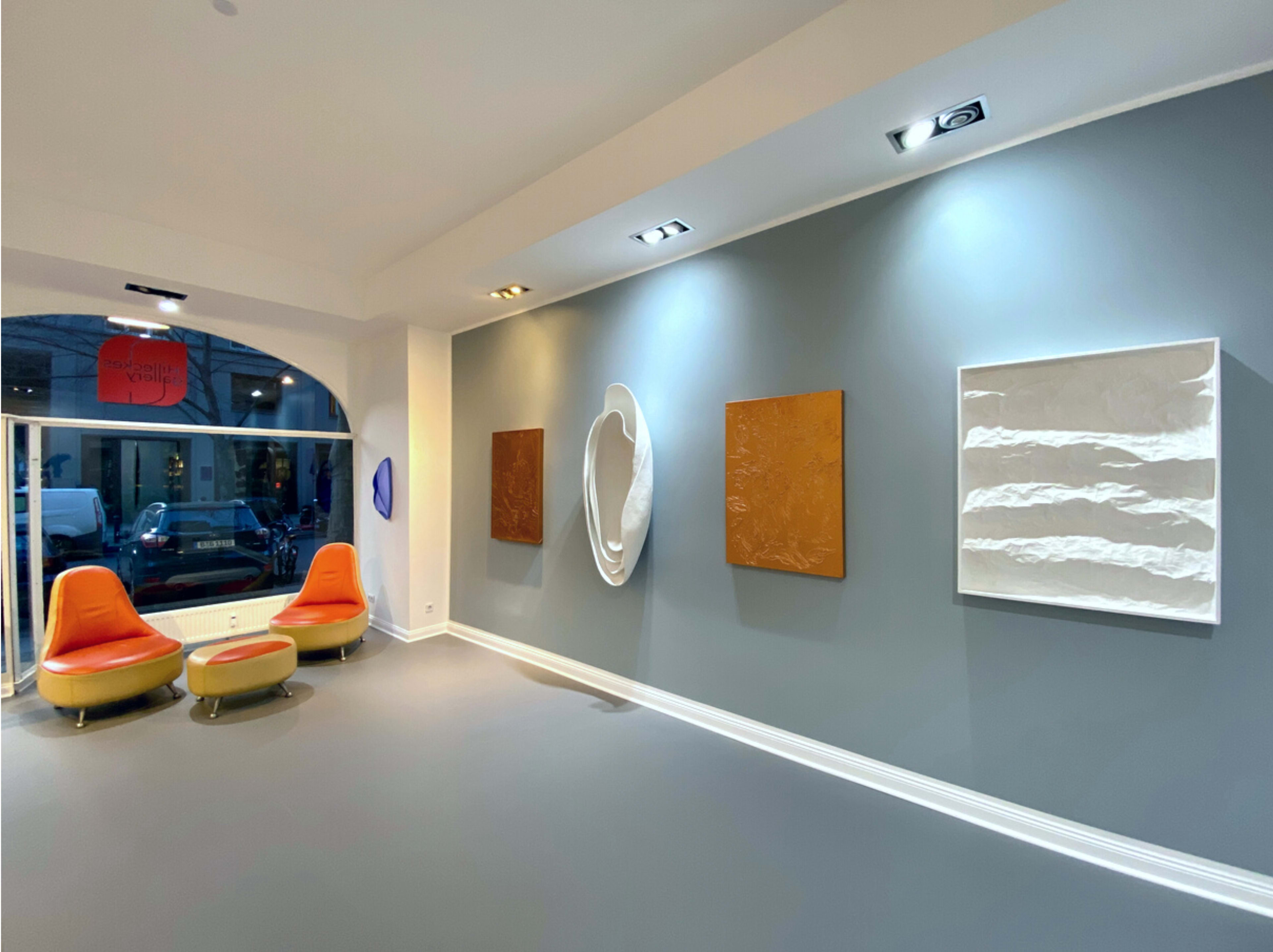 An art gallery interior featuring two orange chairs, three textured wall artworks, and a window displaying street views.