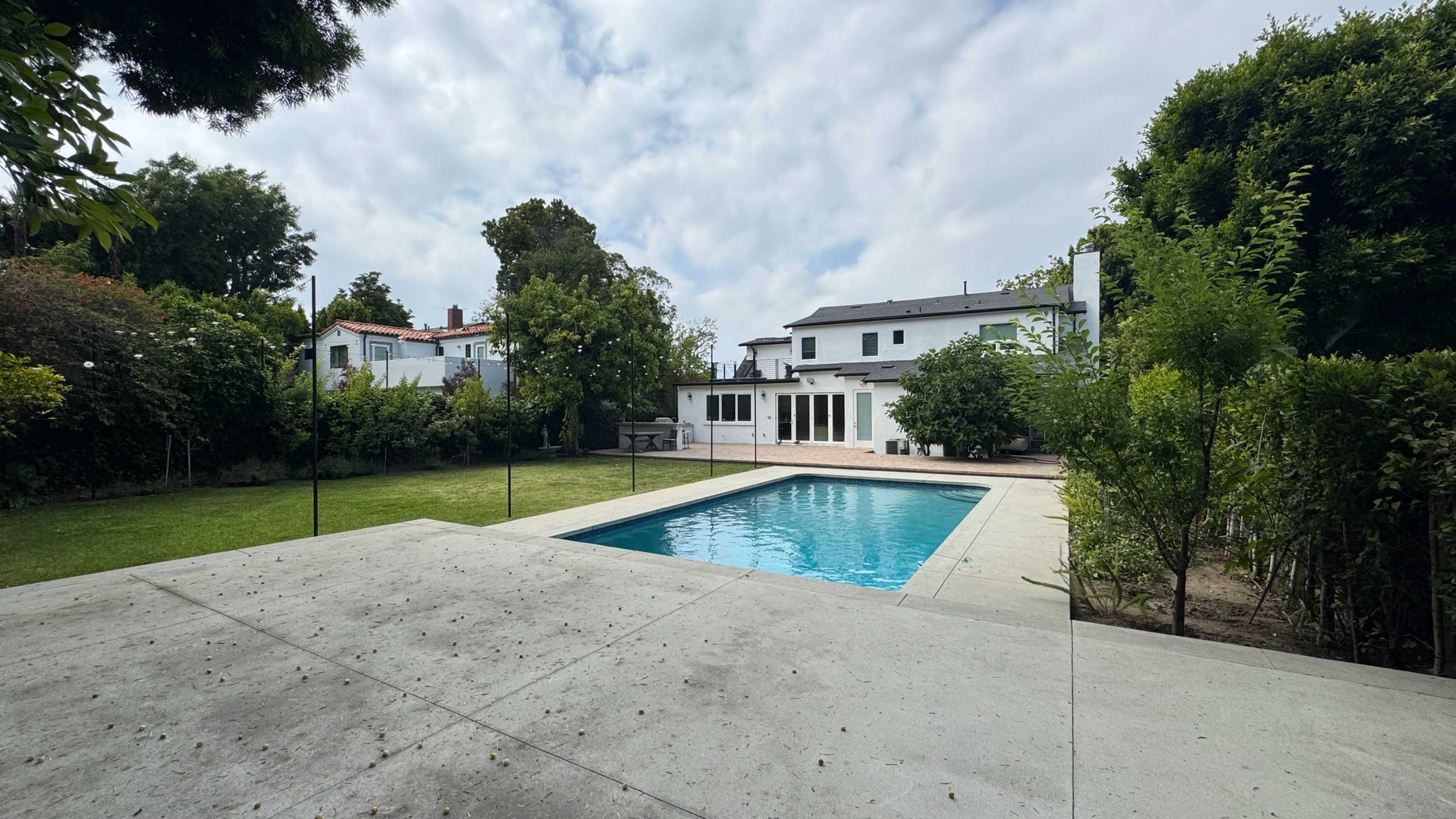 A modern backyard features a swimming pool surrounded by a concrete patio, with a green lawn and trees in the background.