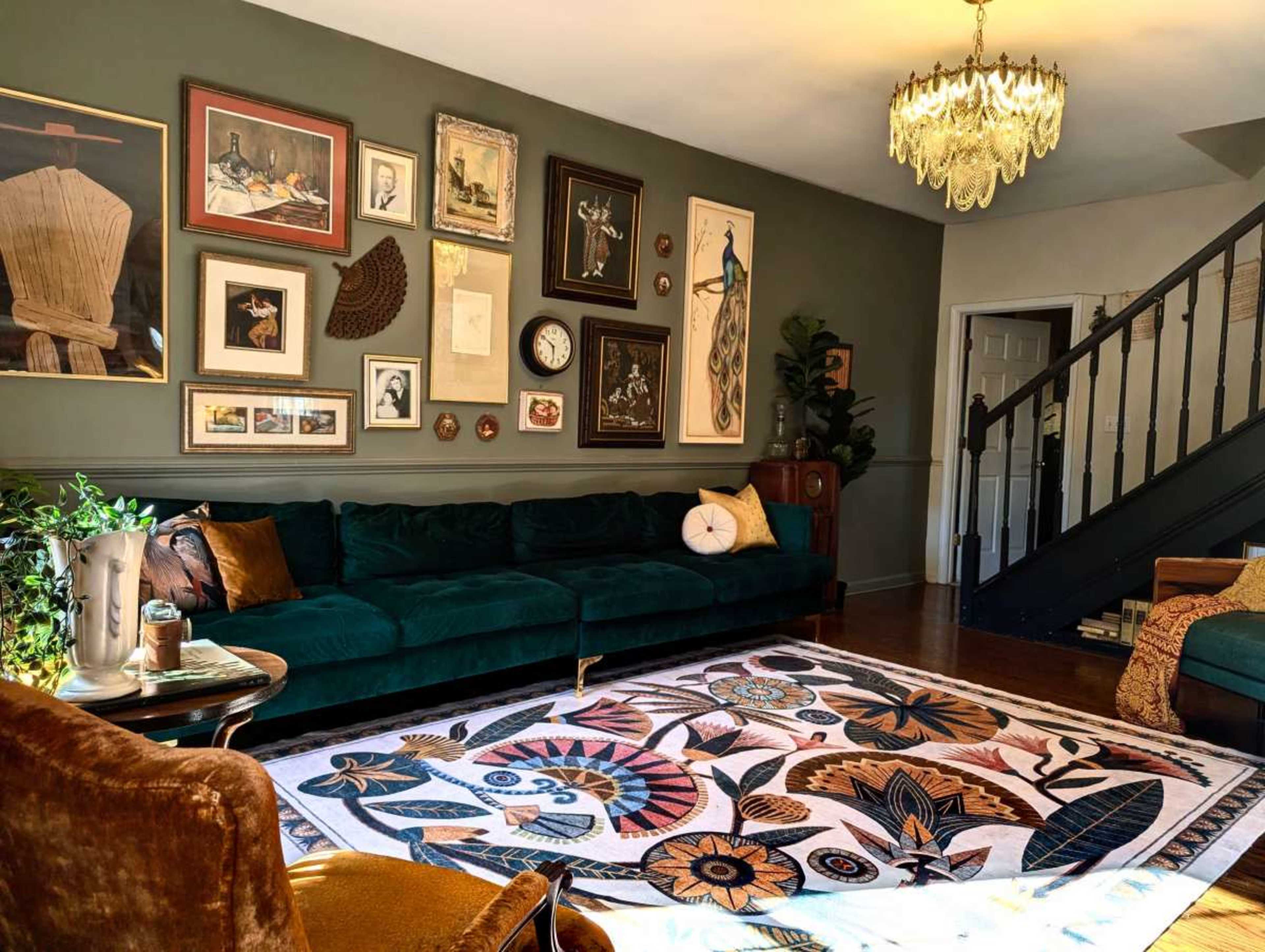 The image shows a cozy living room with a green velvet sofa, various wall art pieces, and a patterned area rug.