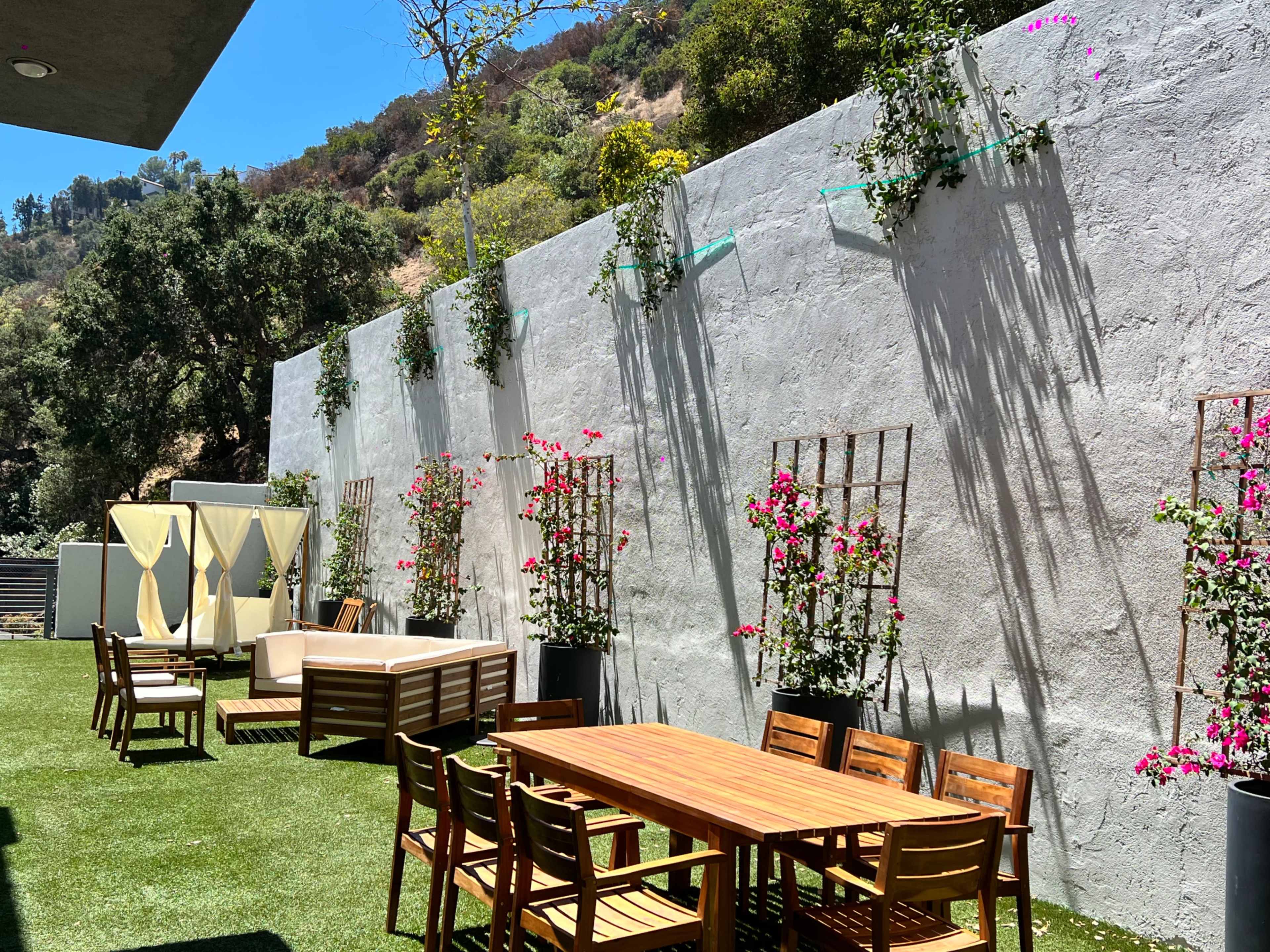 A landscaped outdoor patio featuring a wooden dining table surrounded by chairs, a lounging area with sofas, and vertical planters with pink flowers against a textured wall.