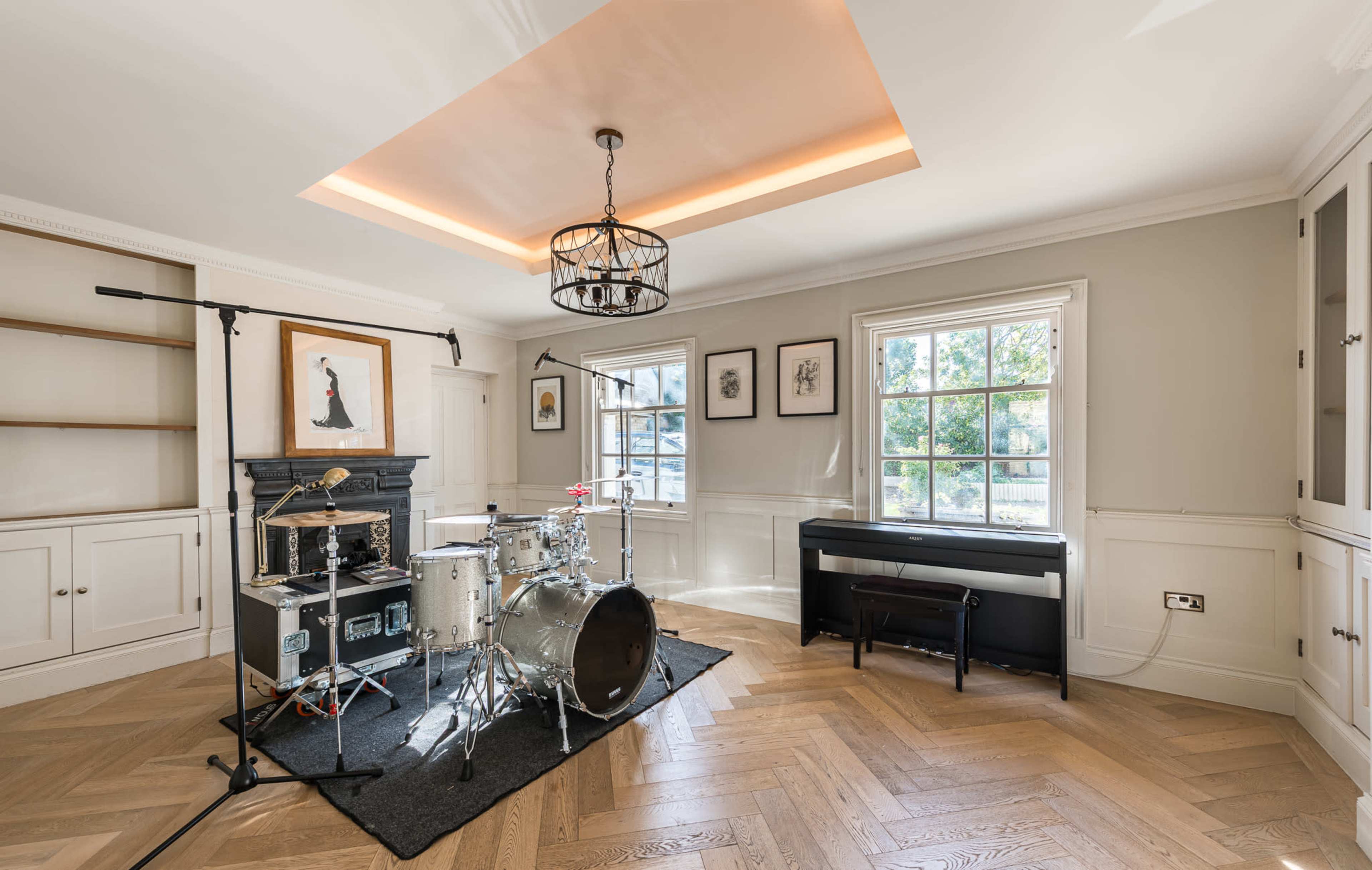 The image shows a well-lit room featuring a drum set on a black mat, a piano, and a shelf area, with large windows letting in natural light.