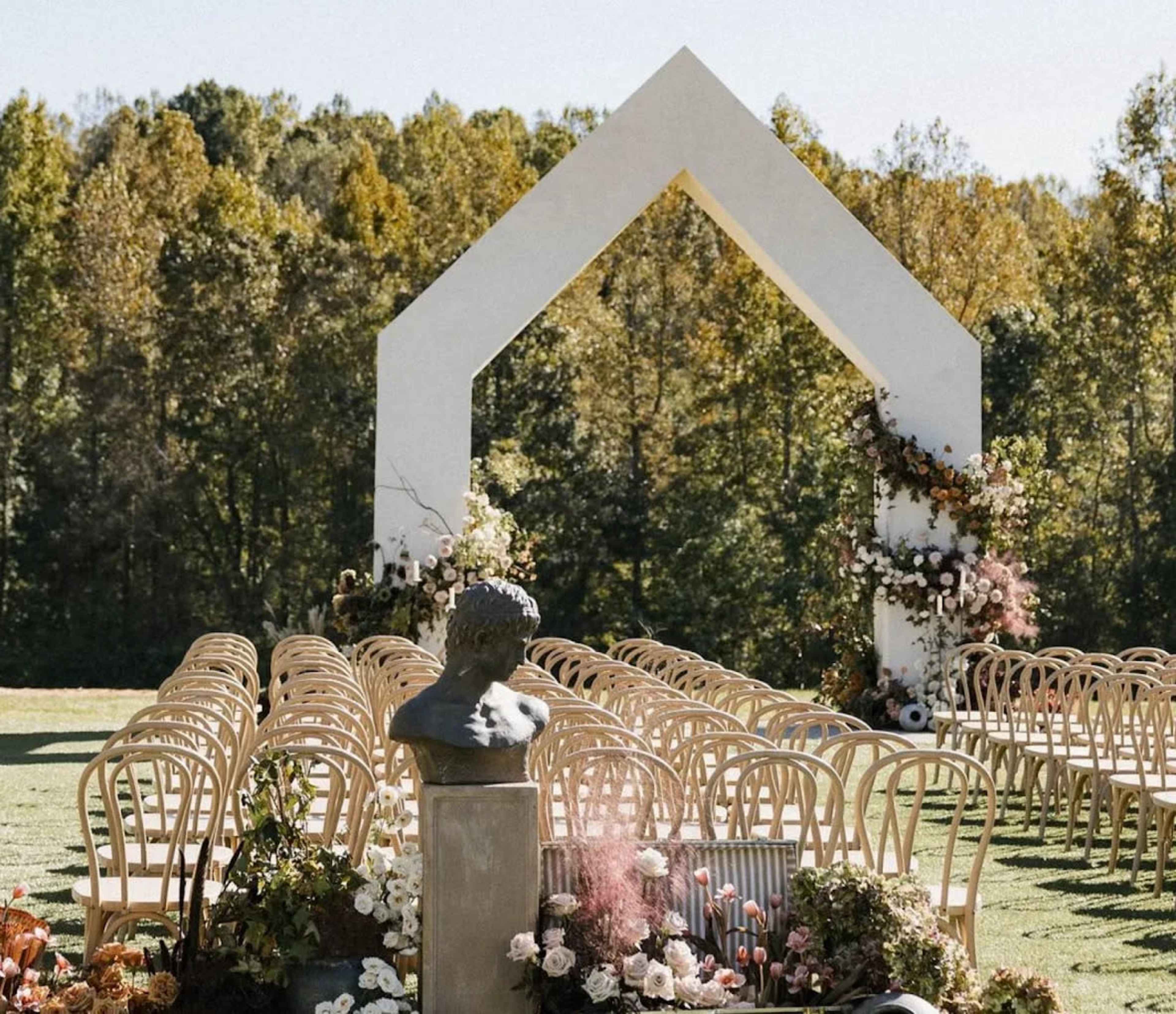 An outdoor wedding ceremony setup features a large white arch adorned with flowers, facing rows of wooden chairs and a bust statue surrounded by floral arrangements.
