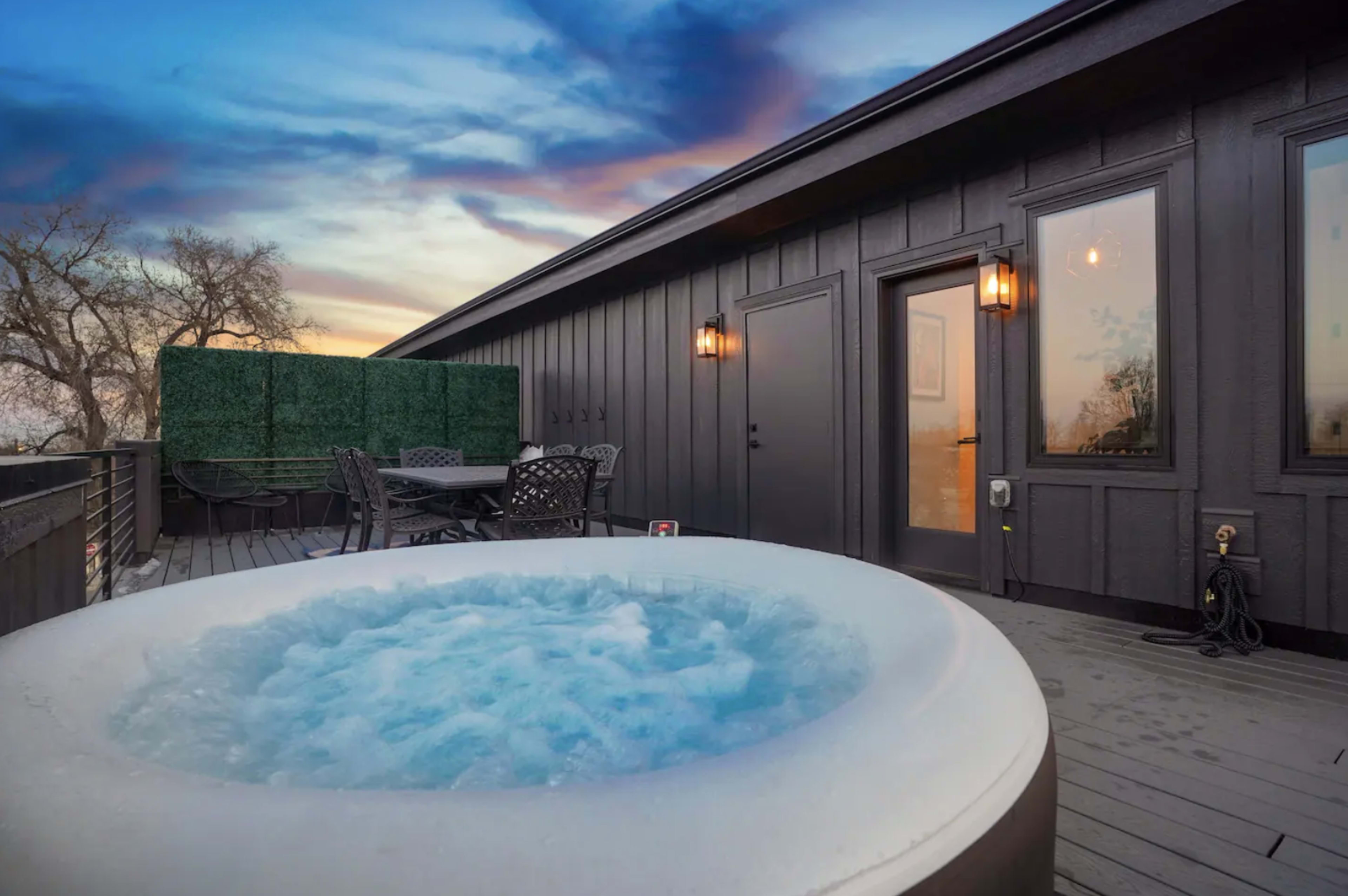 A rooftop deck features a hot tub surrounded by outdoor seating and a backdrop of colorful evening skies.