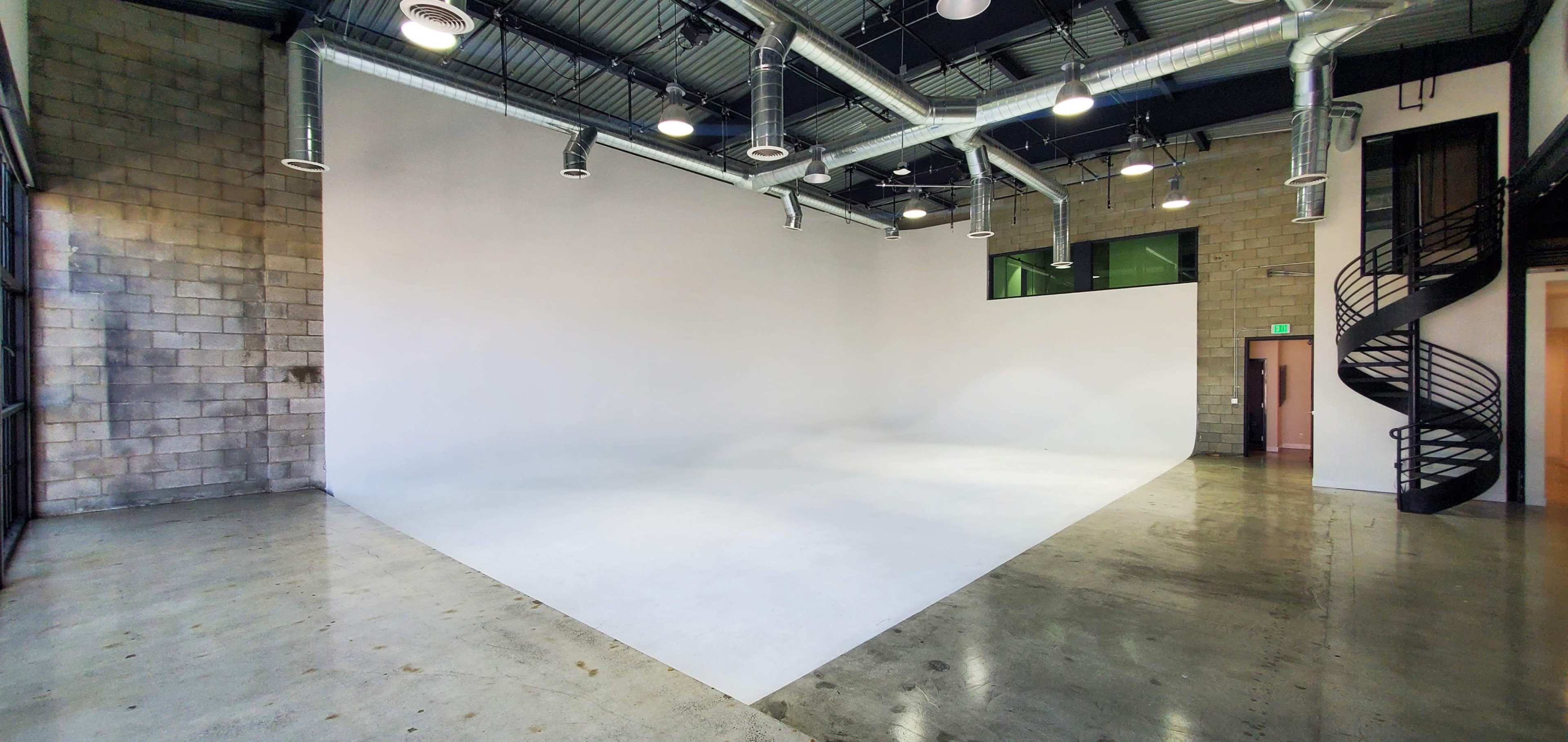A spacious, empty studio with a white backdrop and polished concrete floors, featuring large windows and a spiral staircase.