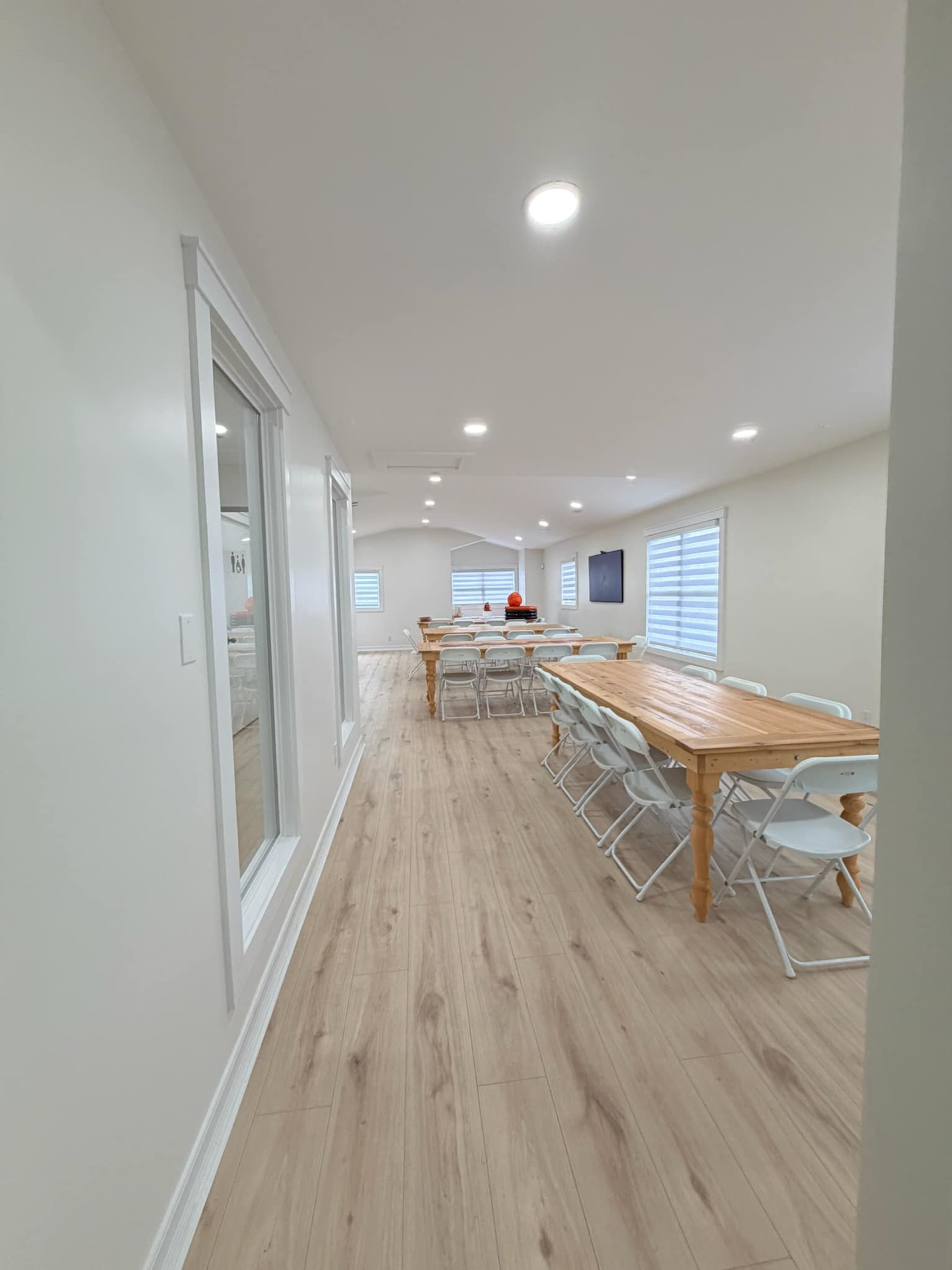 The image shows a long, narrow room with a wooden dining table surrounded by chairs, and bright white walls with large windows.