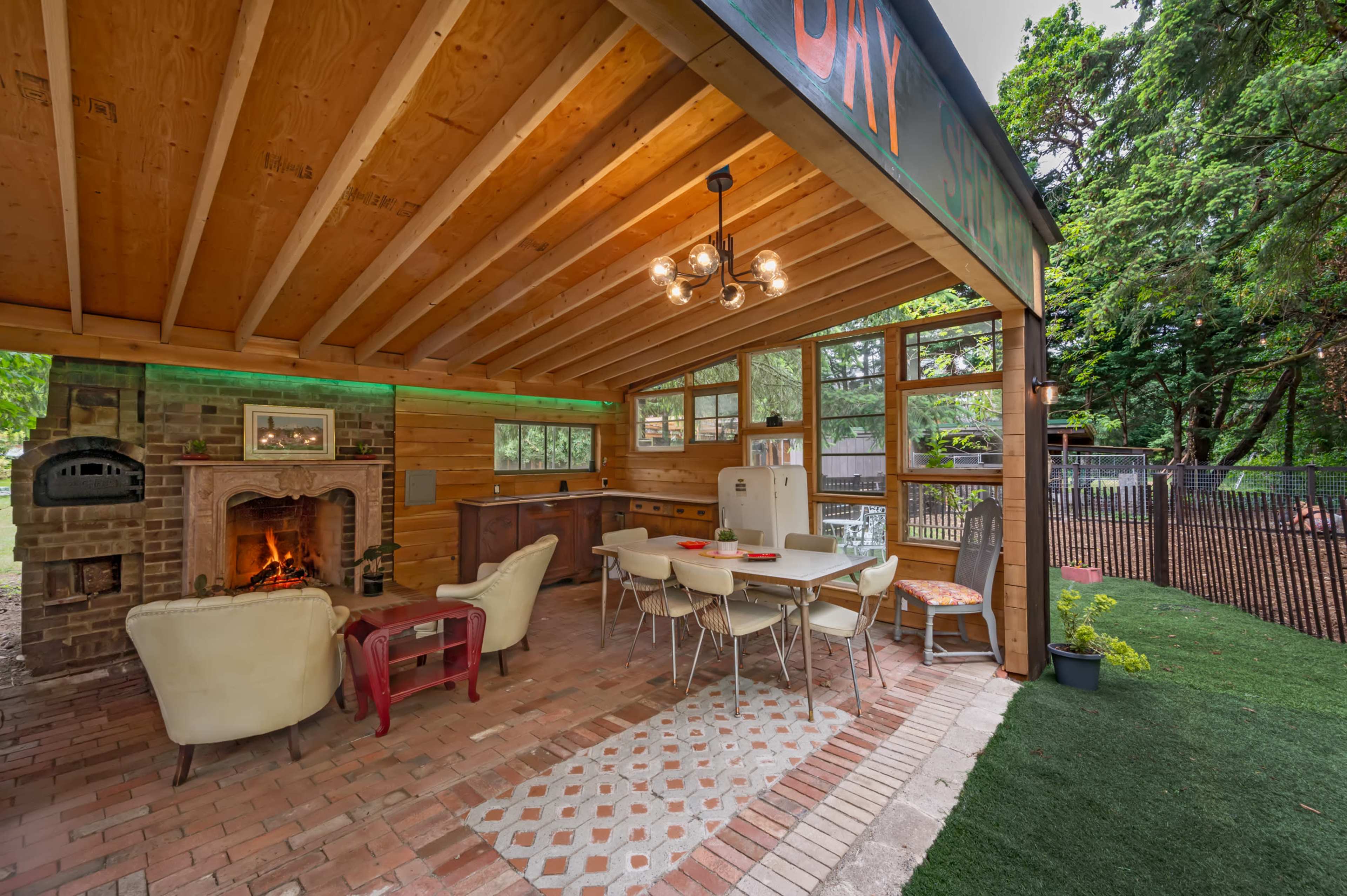 A rustic outdoor living space featuring a dining area, a brick fireplace, and large windows with a view of a lush green garden.