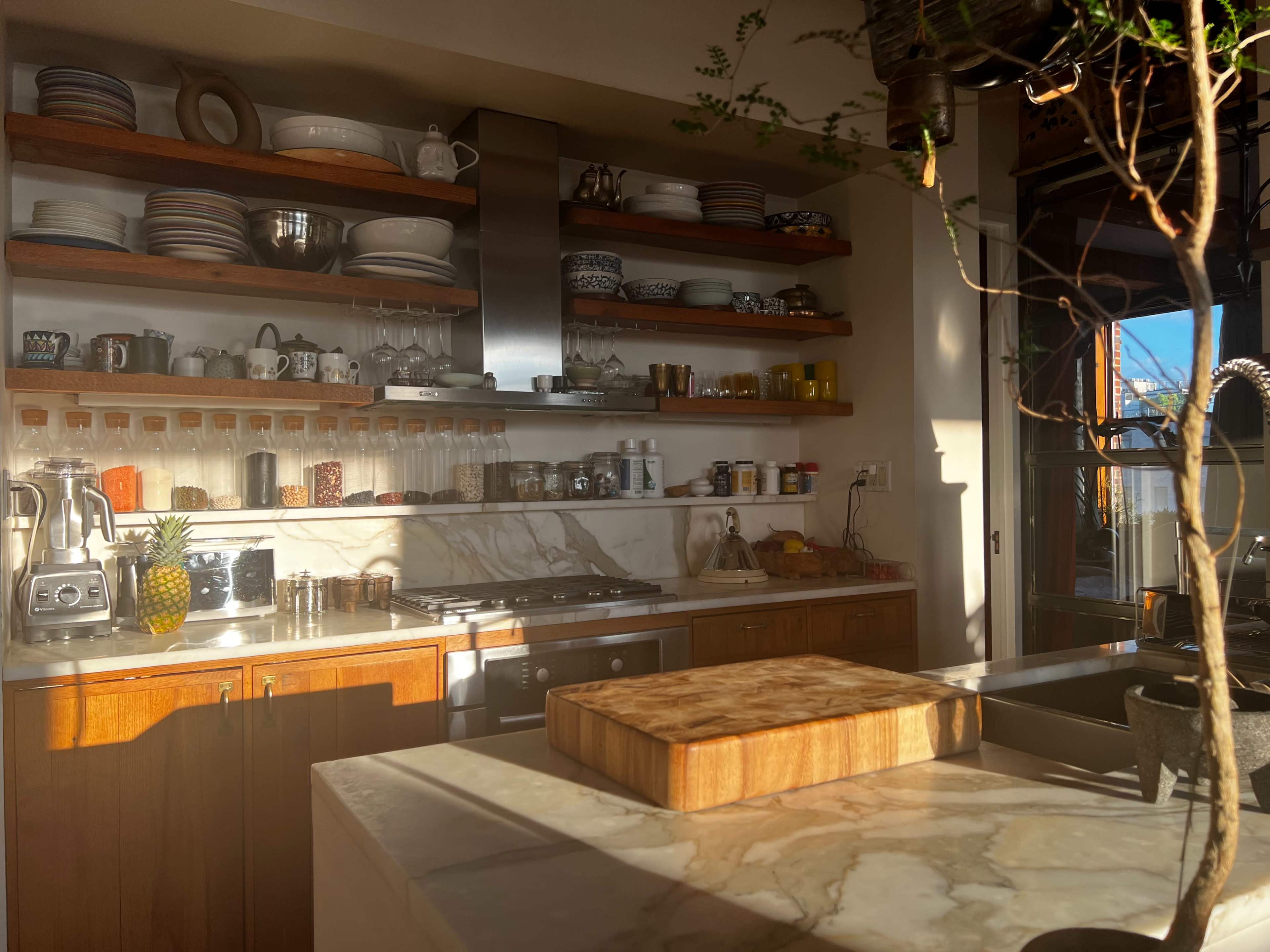 The image shows a modern kitchen with open shelves displaying dishes and glassware, a marble countertop, and a stovetop with a cutting board.