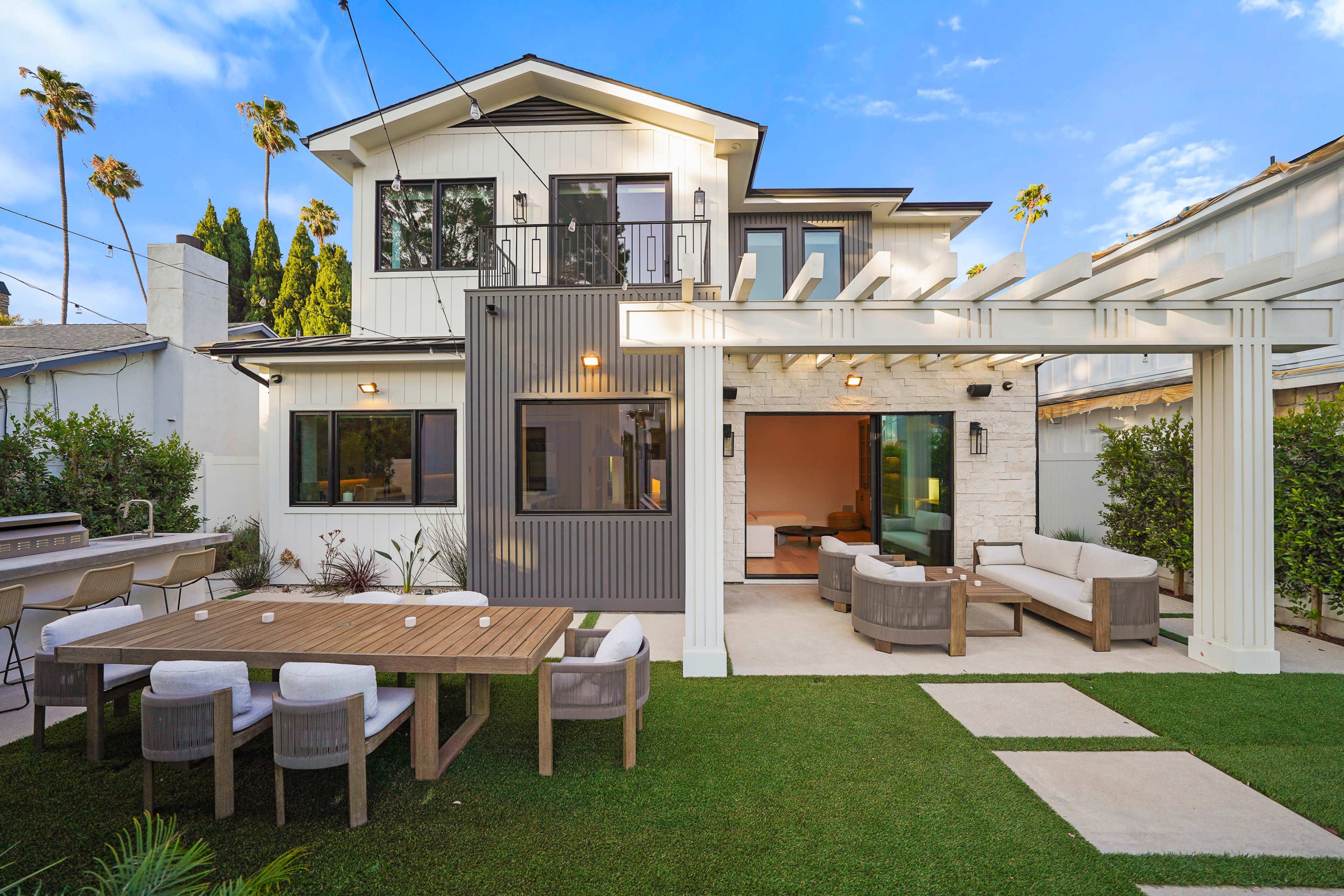 The image shows a modern home with a patio featuring outdoor seating and a dining table, surrounded by greenery and palm trees.