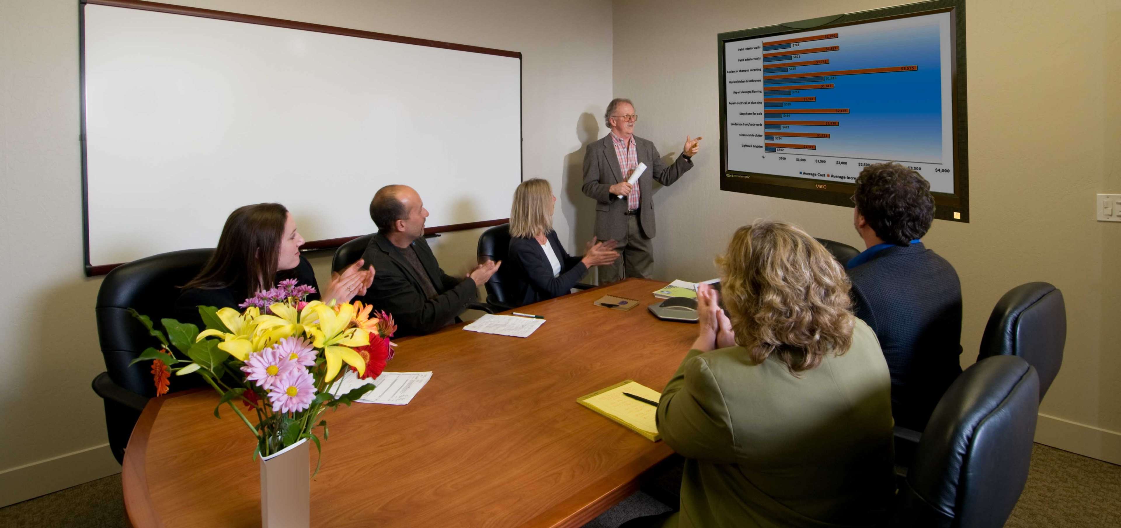 A group of six professionals is seated around a conference table, watching a presentation with a chart displayed on a large screen.