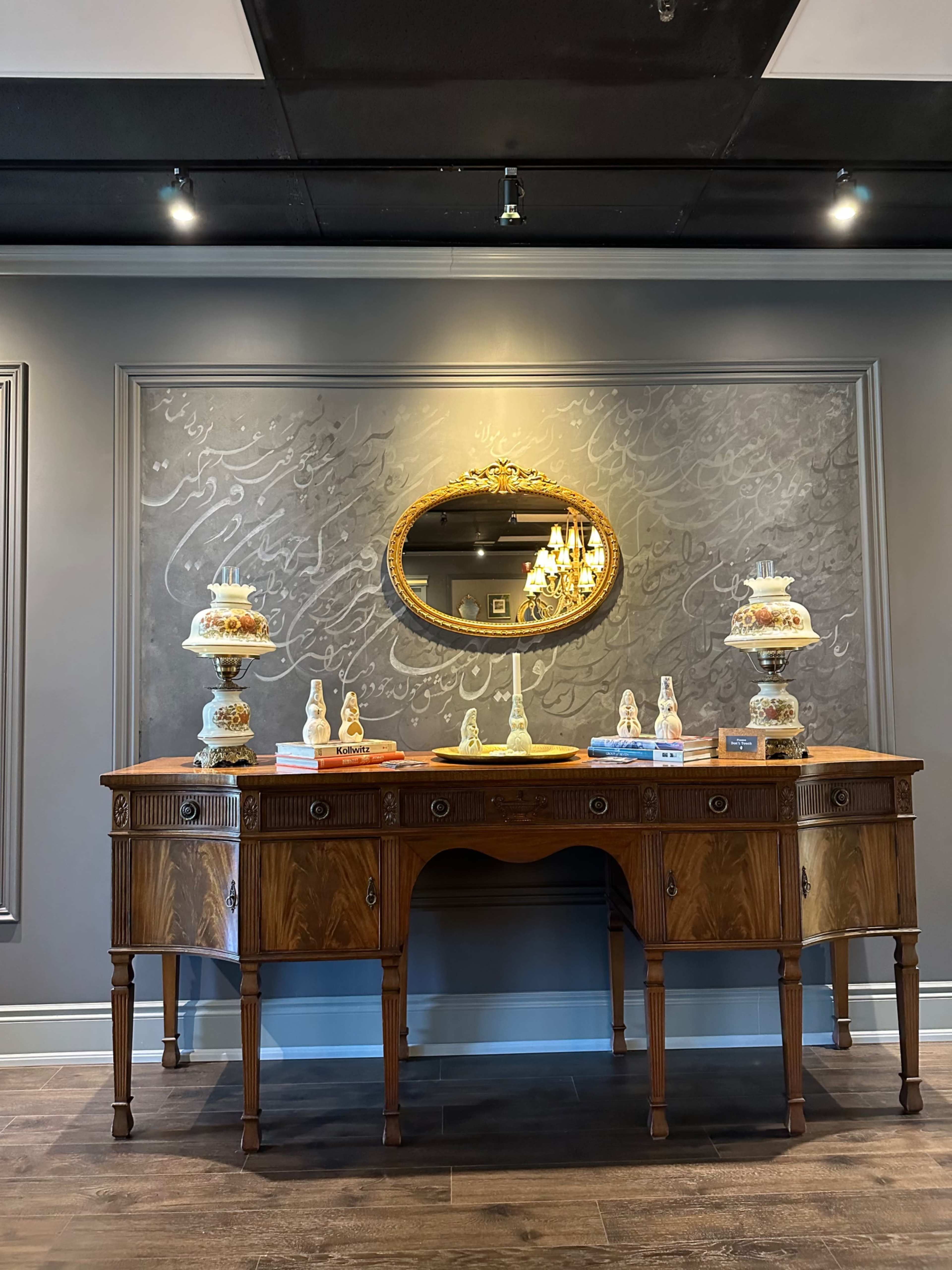 A wooden sideboard with decorative cakes on stands is placed against a wall featuring ornate calligraphy and a framed mirror.