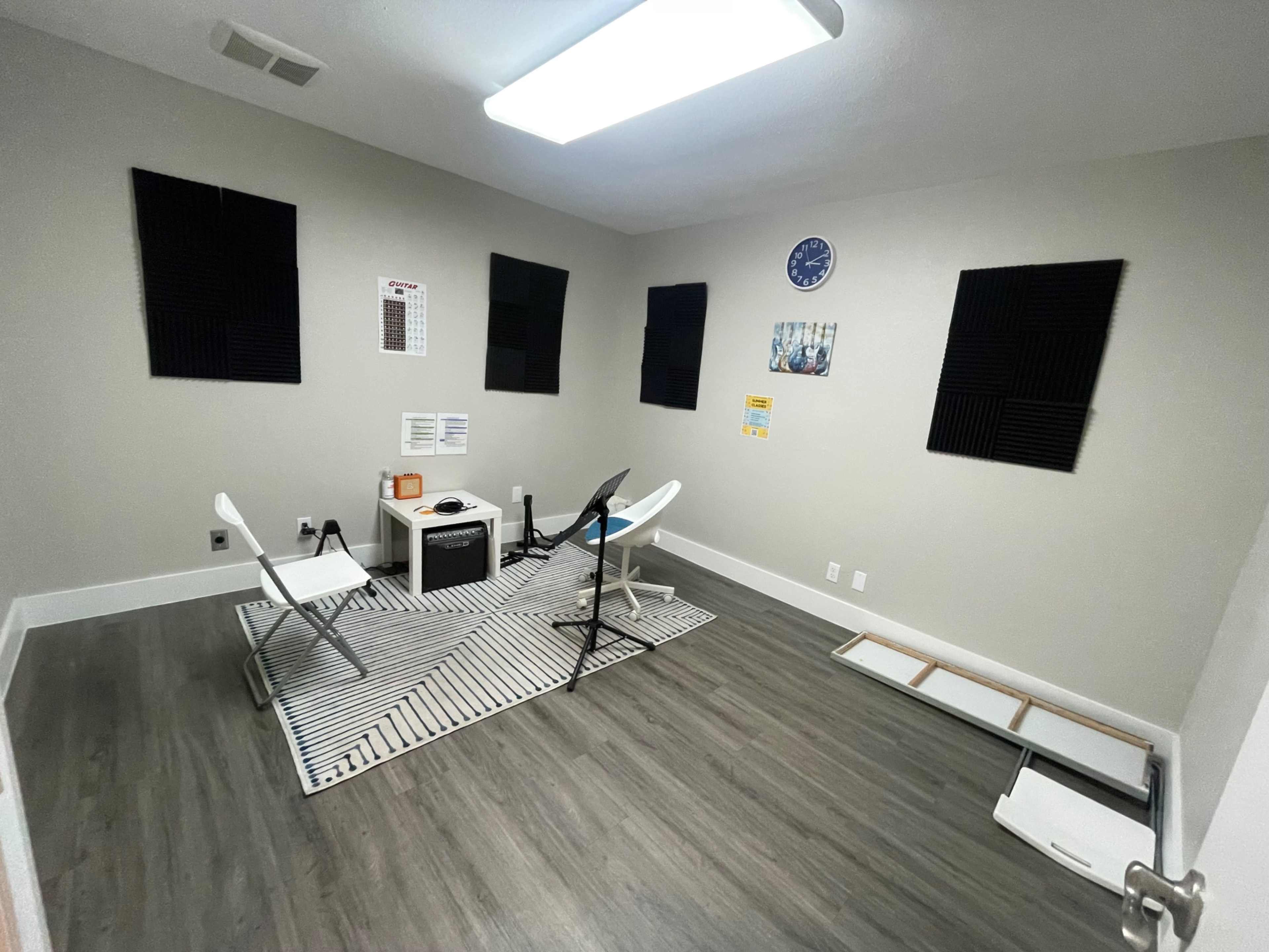 The room features gray walls, a patterned rug, two white chairs, a small table, acoustic foam panels on the walls, and a wall clock.