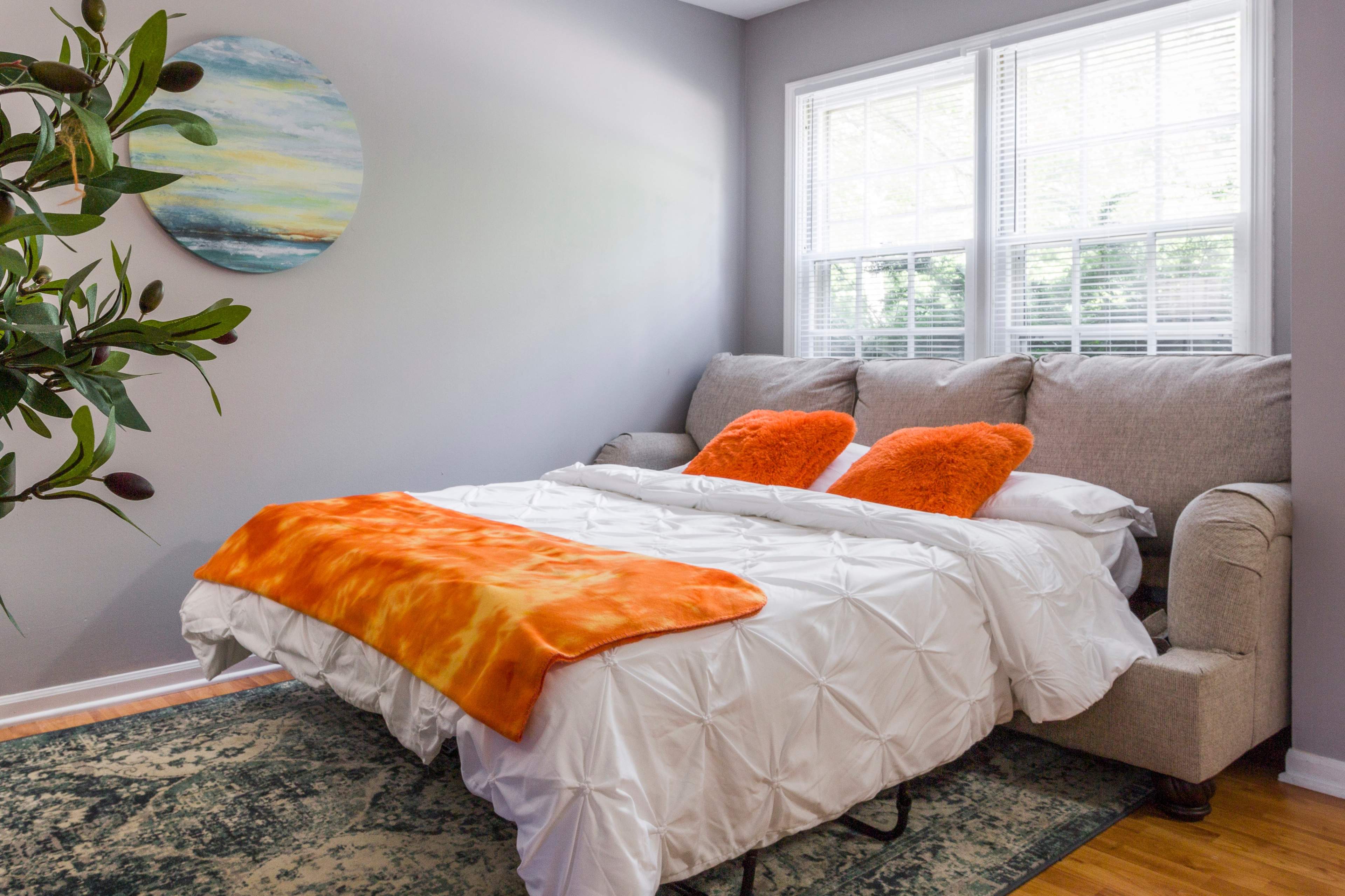 A small bedroom features a sofa that converts into a bed, topped with an orange blanket and two orange cushions, next to a plant and large windows.