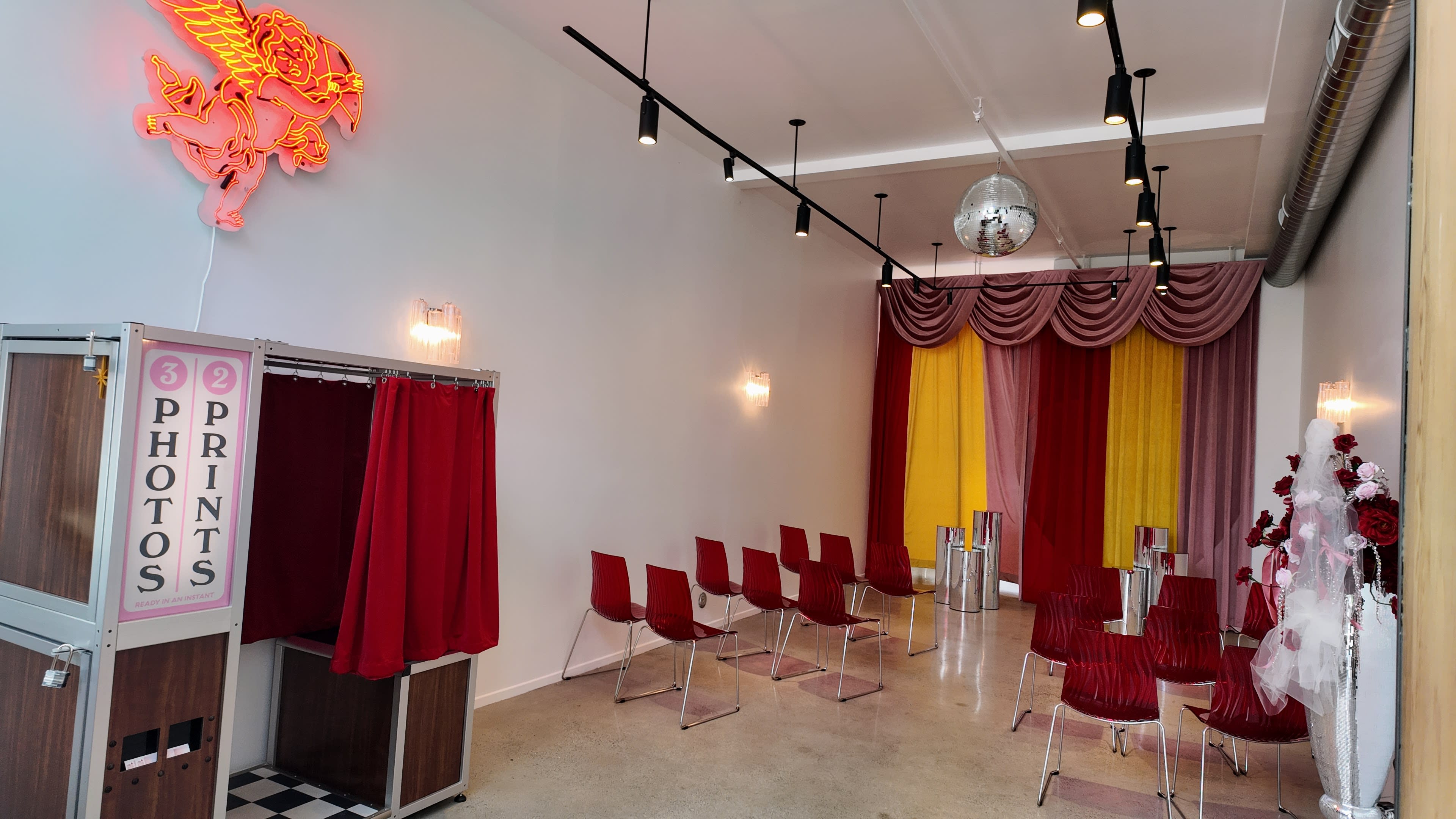A brightly lit interior space featuring a photo booth, red curtains, colorful fabrics, and several red chairs arranged in a waiting area.