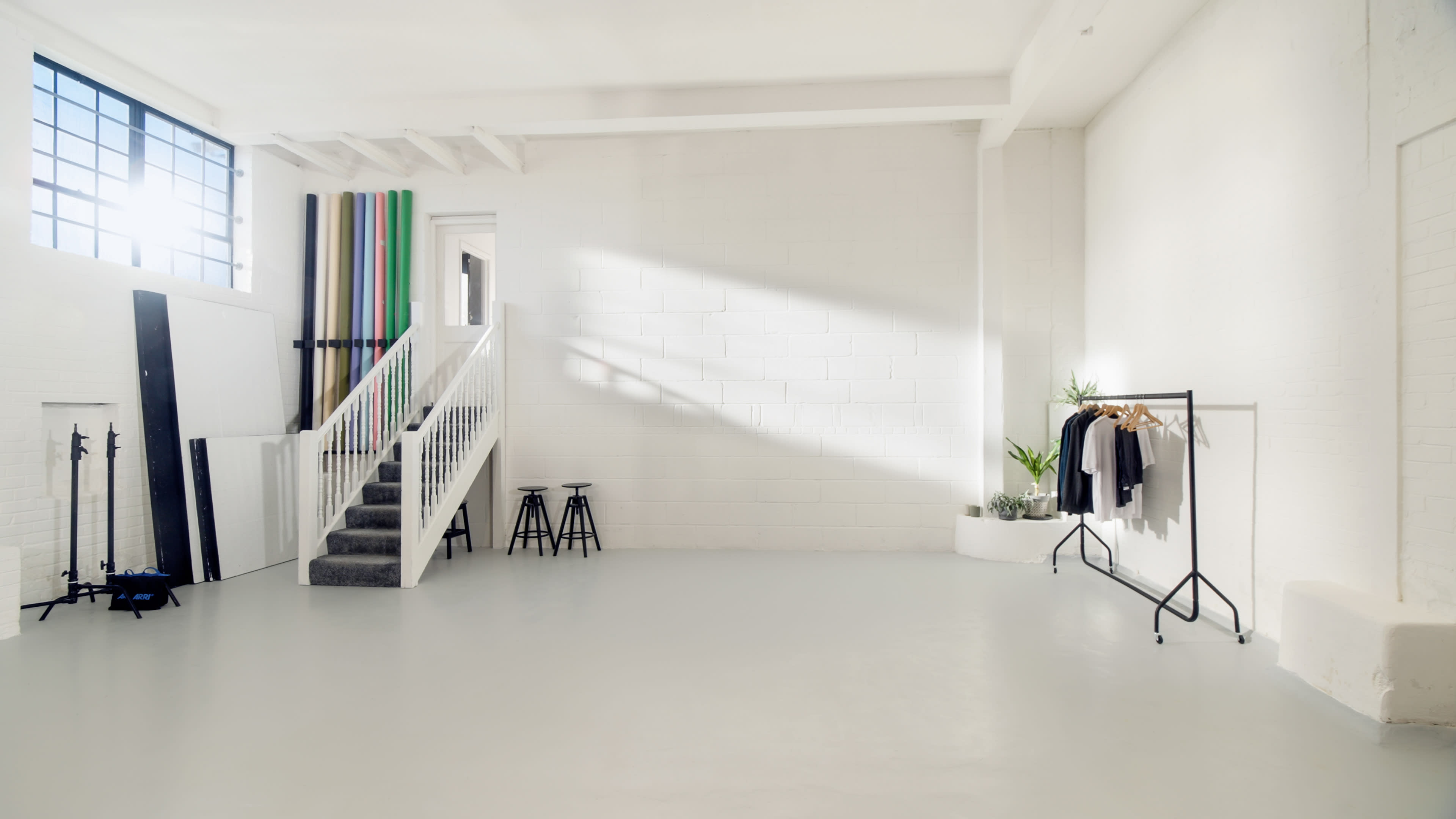 The image shows an empty, well-lit studio space featuring a staircase, a clothing rack with garments, and backdrop rolls in various colors.