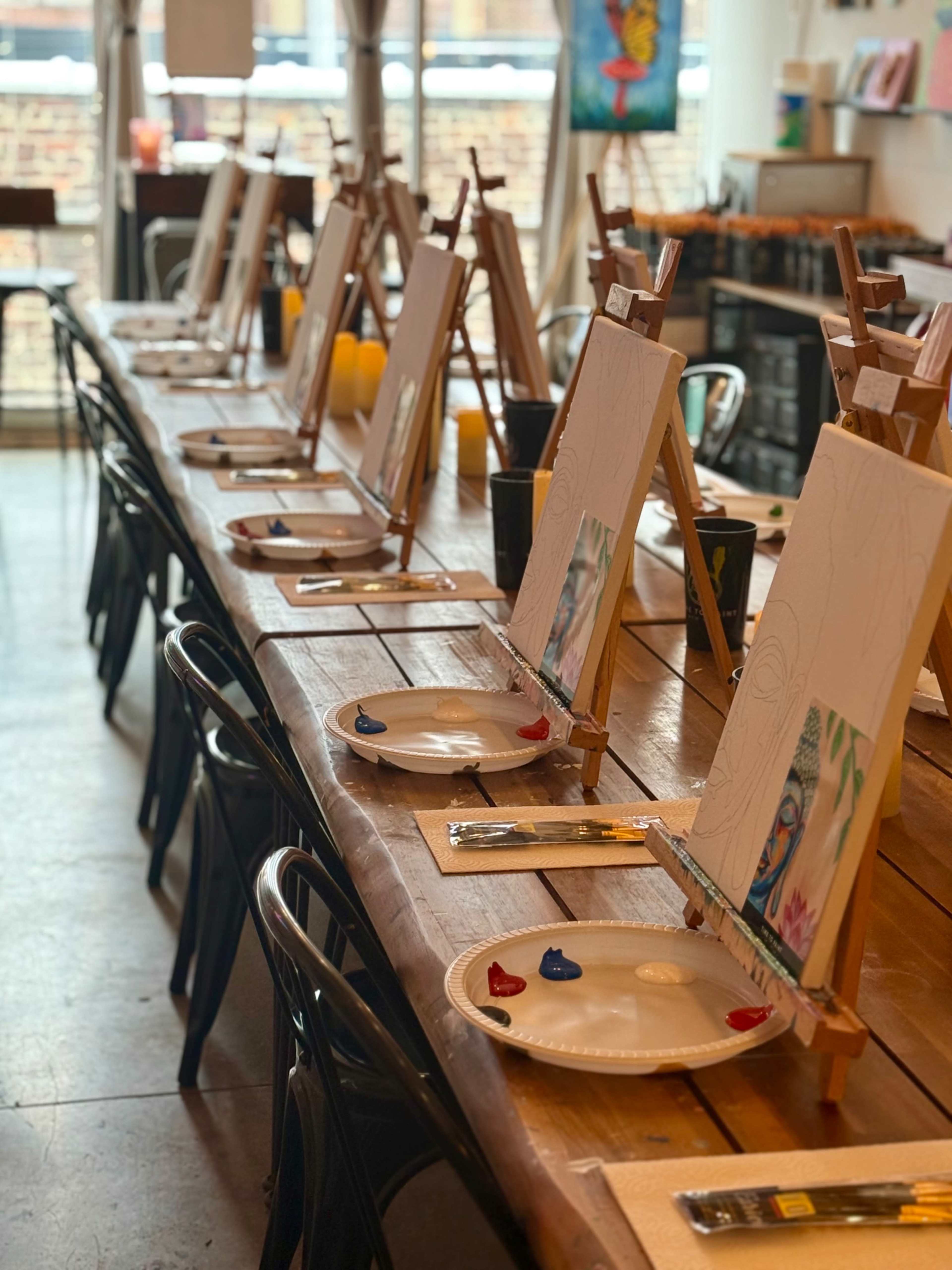 The image shows a long wooden table set up for a painting class, with easels displaying blank canvases and palettes with paint colors at each place setting.