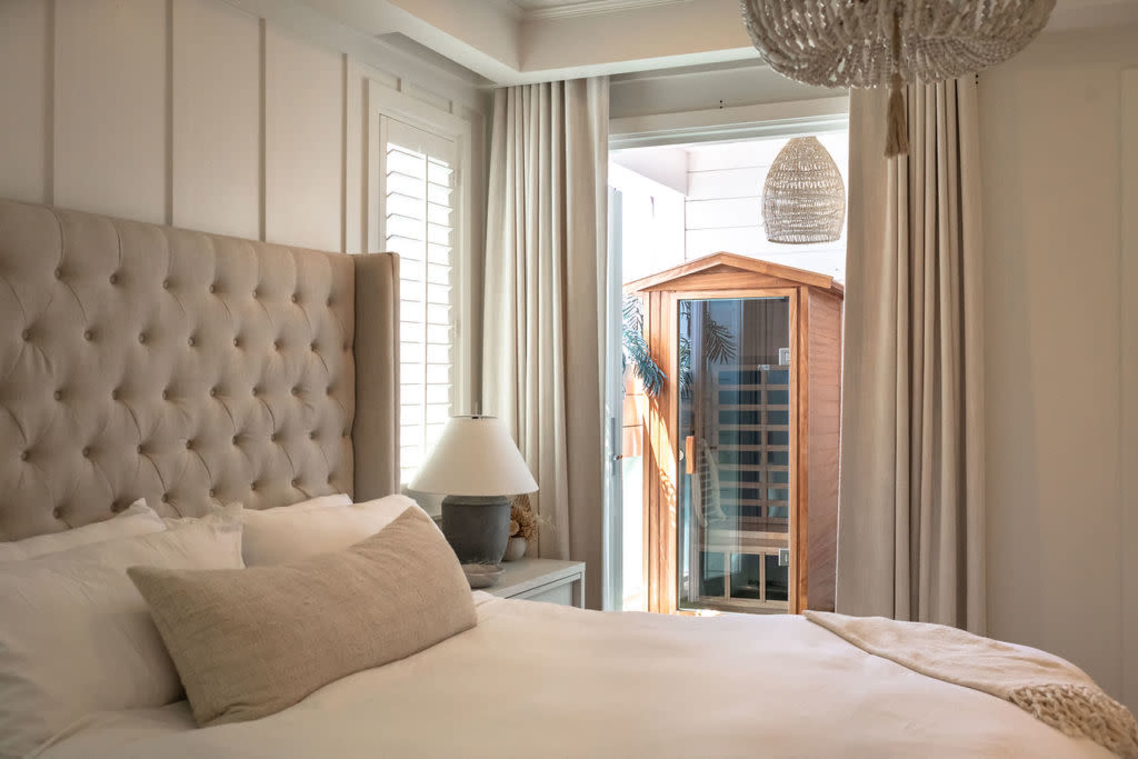 A well-decorated bedroom features a large upholstered headboard, a bedside lamp, and a view of a wooden sauna through an open door.