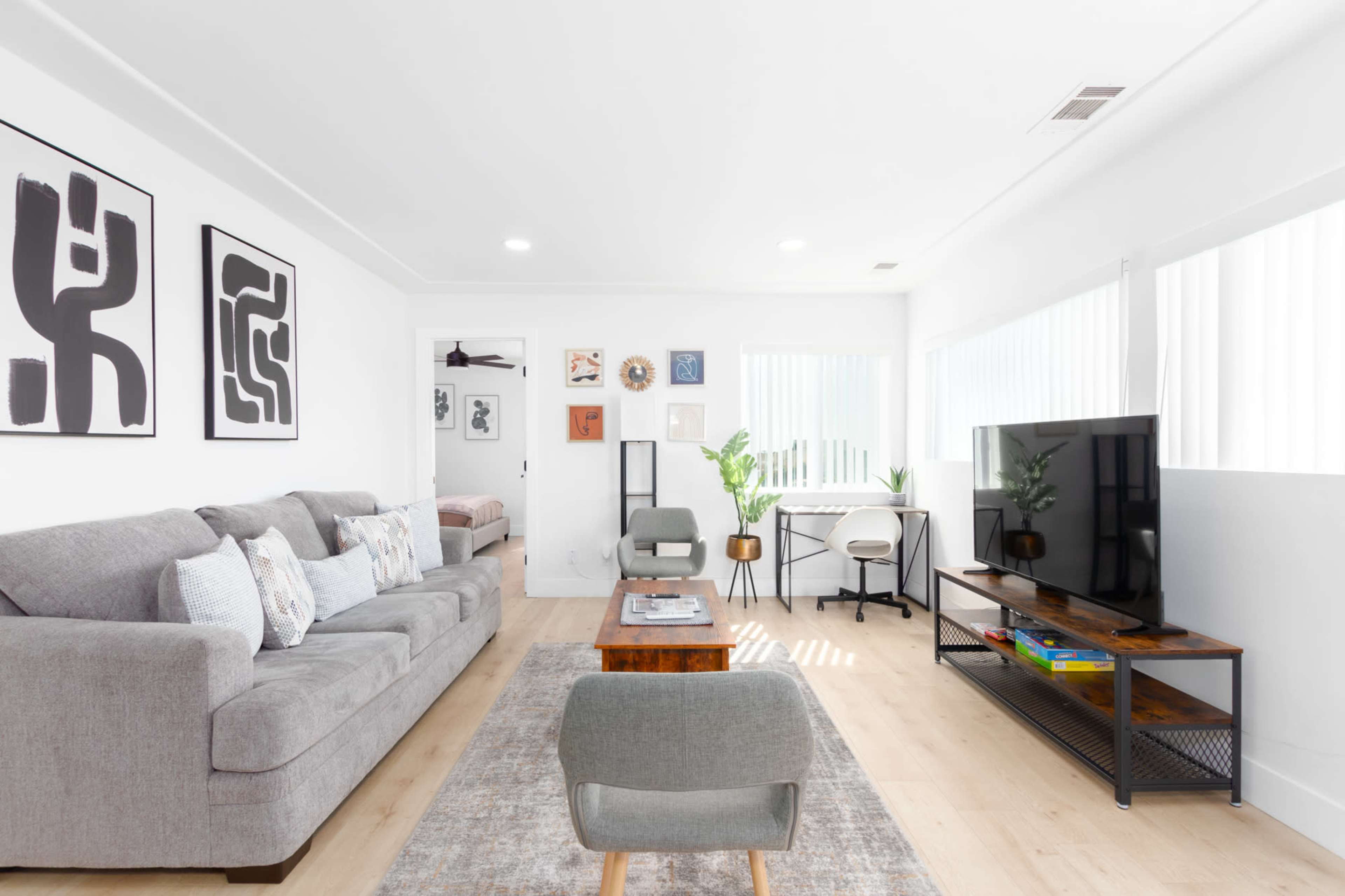 A modern living room features a gray sectional sofa, a wooden coffee table, a mounted TV, and large windows with blinds, framed by abstract wall art.