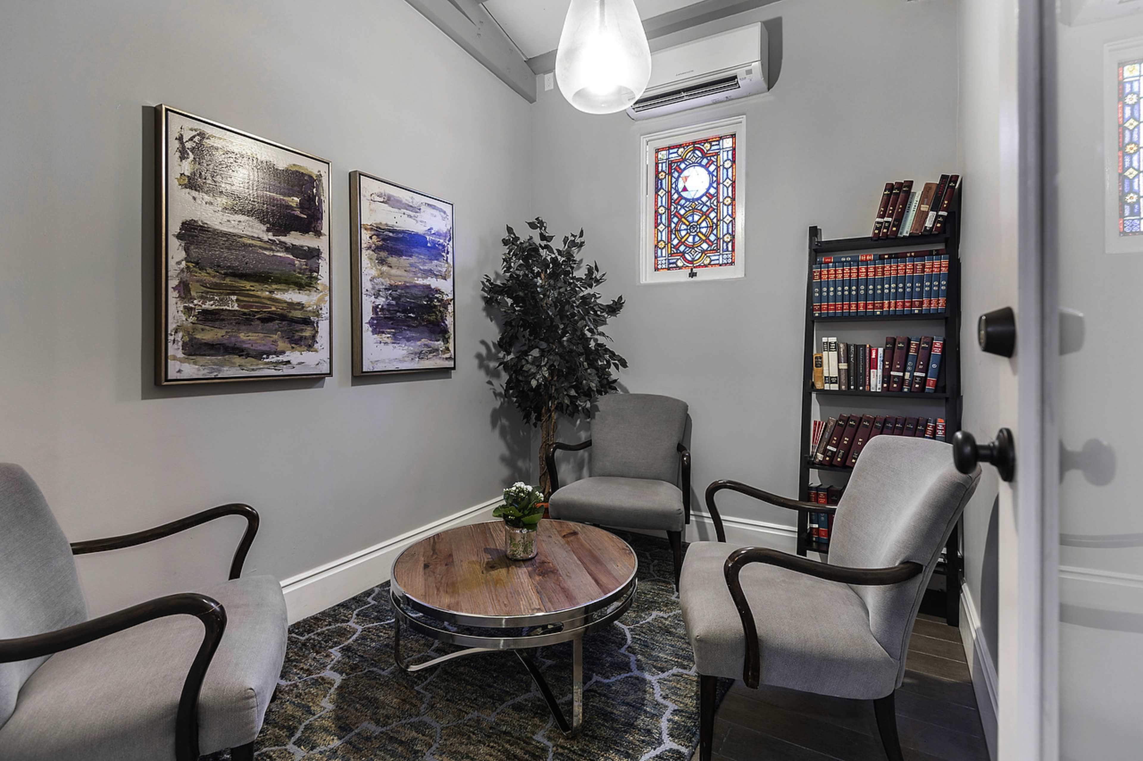 A small, well-furnished room features two chairs, a round wooden table, a potted plant, a bookshelf with books, and two framed artworks on the walls.