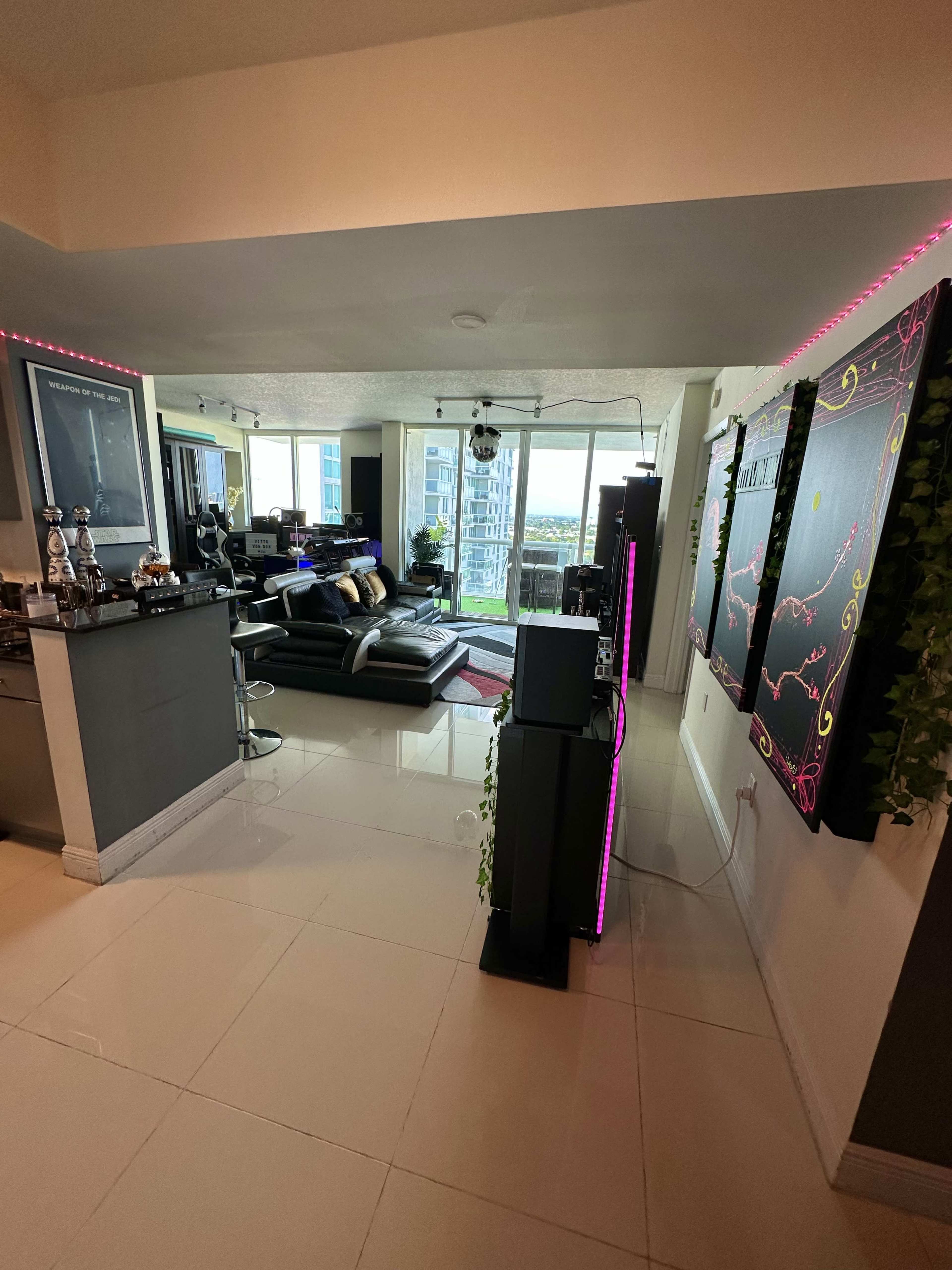 A modern living space with a view of city buildings, featuring a kitchen area on the left and a black leather sectional sofa in the sitting area.