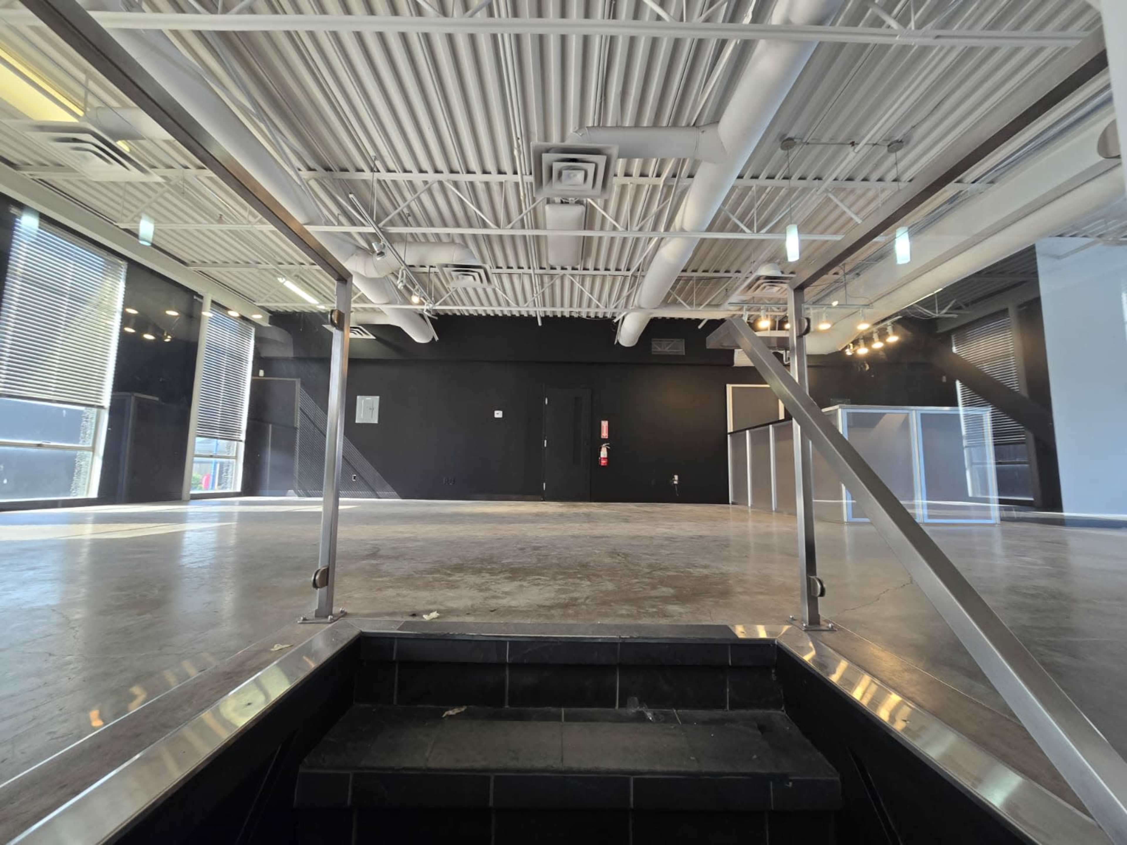 The image shows a large, vacant commercial space with a staircase leading down to the floor, illuminated by natural light coming through large windows.