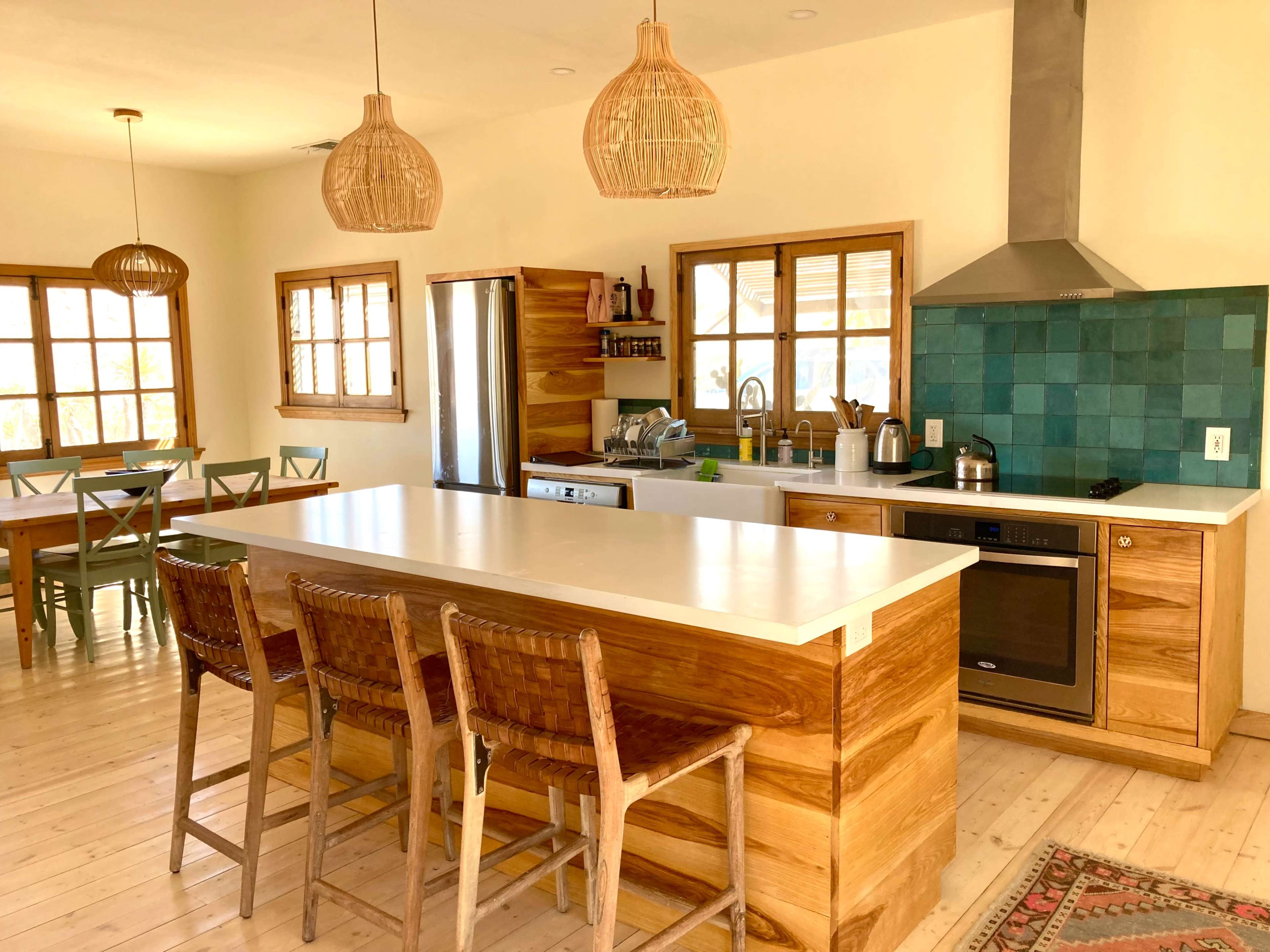 A modern kitchen features wooden cabinetry, a large island with seating, and a teal tile backsplash, with a dining area visible in the background.